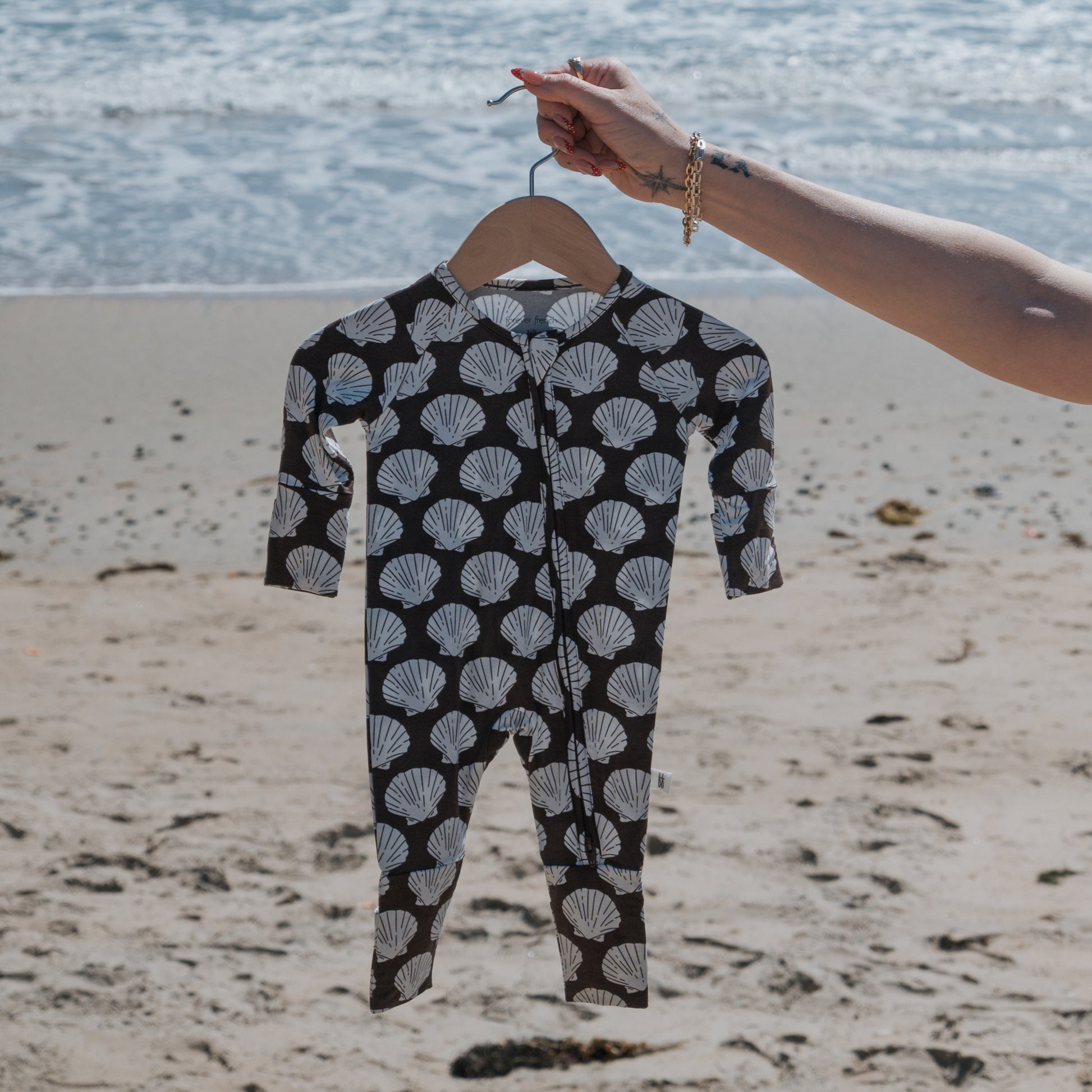 A hand holds forever french baby’s Bamboo Zip Pajamas | Seashells—a black baby romper with a white seashell pattern—made from hypoallergenic fabric, displayed in front of a sandy beach and ocean waves.
