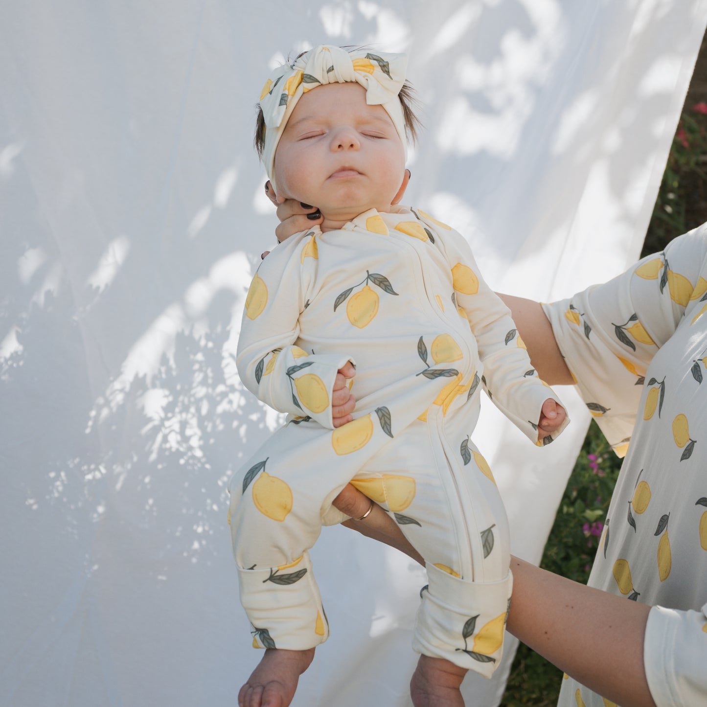 An adult gently holds a baby wearing forever french baby's Bamboo Zip Pajamas | Lemon with a matching headband. Sunlight and shadows on a white sheet outdoors highlight the breathable comfort and lemon-themed style.
