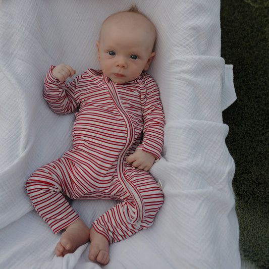 A baby in forever french baby Bamboo Zip Pajamas | Heritage Stripe—a red and white striped onesie—lies on a white blanket on green grass, gazing up with one hand slightly raised.