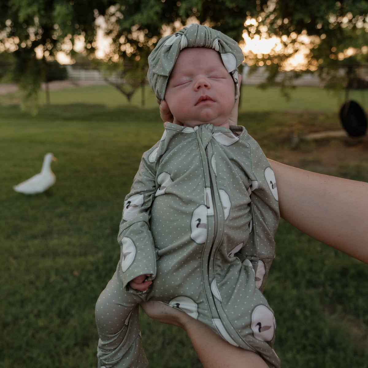 A baby dressed in forever french baby’s Bamboo Zip Pajamas in the Mallard Frames print is held up outdoors at sunset, surrounded by green grass and trees, with a white duck visible in the background.