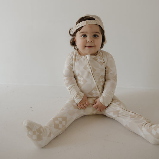 A toddler in forever french baby's Bamboo Zip Pajamas | Cottage Quilt—beige and cream with a geometric pattern—and a backwards beige cap sits on the floor against a white background, looking at the camera with a slight smile.