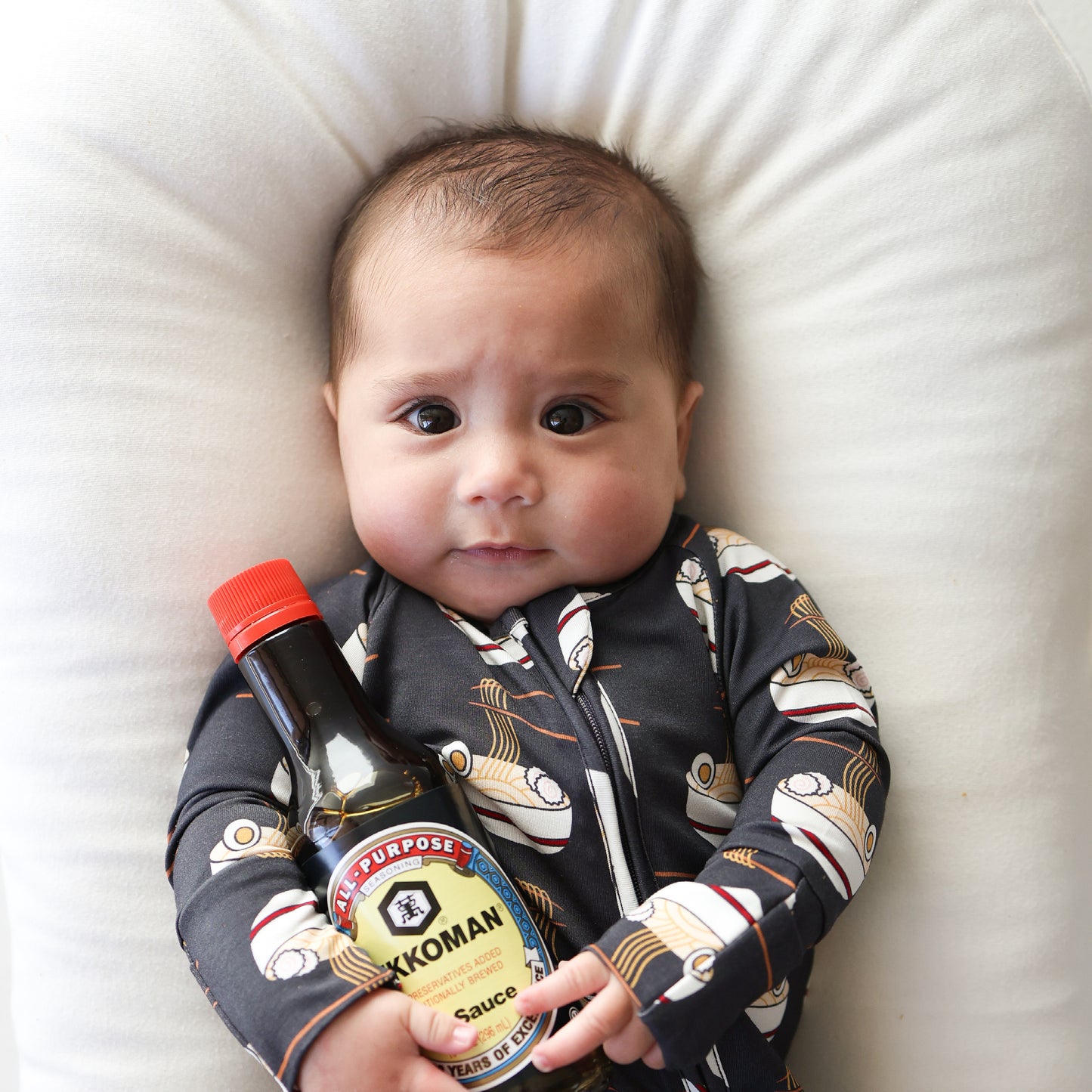 A baby in forever french baby Bamboo Zip Pajamas | Ramen lies on a white cushion, holding a bottle of Kikkoman soy sauce and gazing upward with wide eyes.