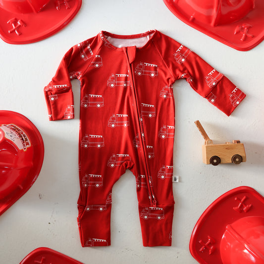 The forever french baby Bamboo Zip Pajamas | Fire Truck are displayed flat on a white background, surrounded by red firefighter hats and a small wooden fire truck toy.