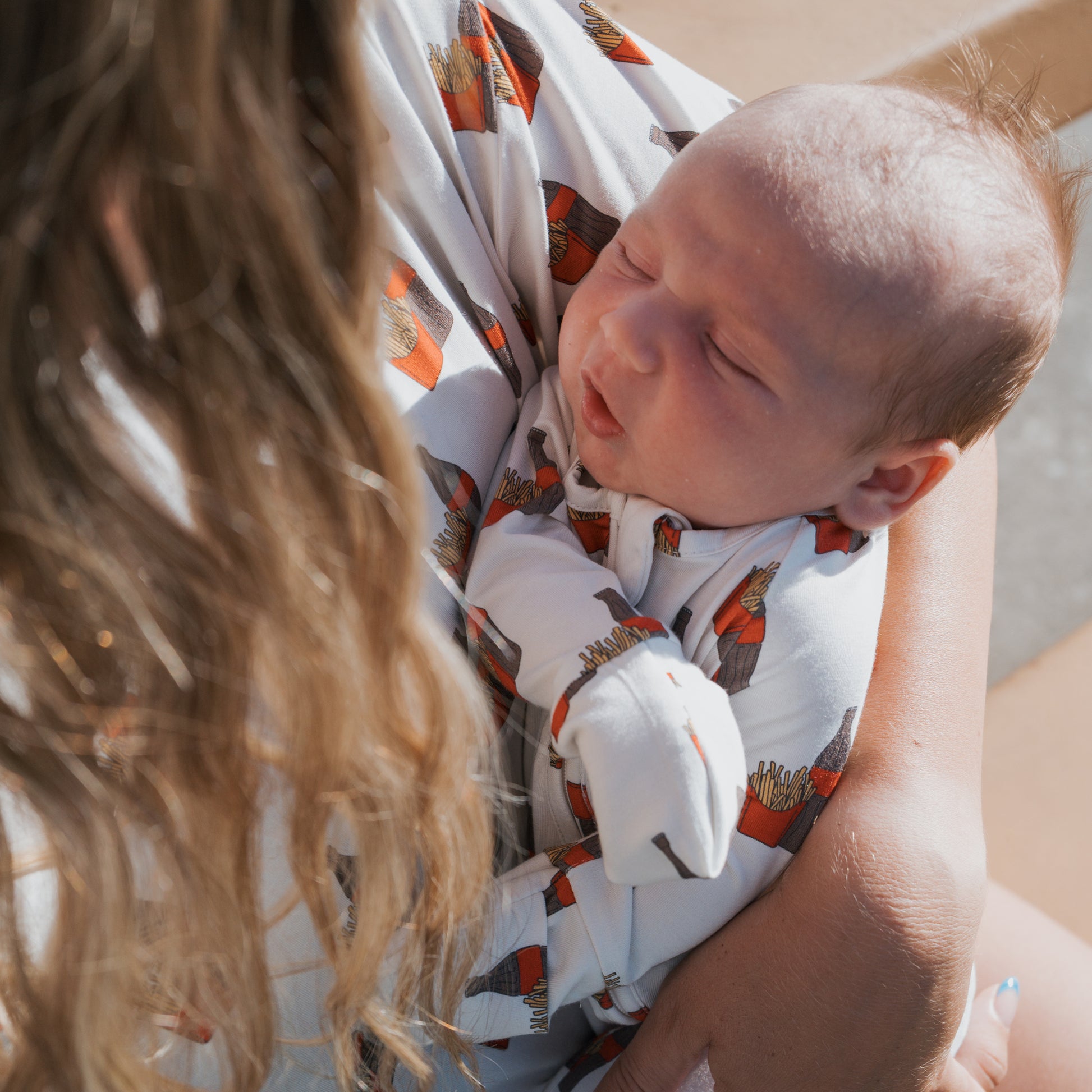 A woman with long, wavy hair holds a sleeping baby dressed in forever french baby's Bamboo Zip Pajamas | Fries & Cola print. The photo is taken outdoors in natural light.