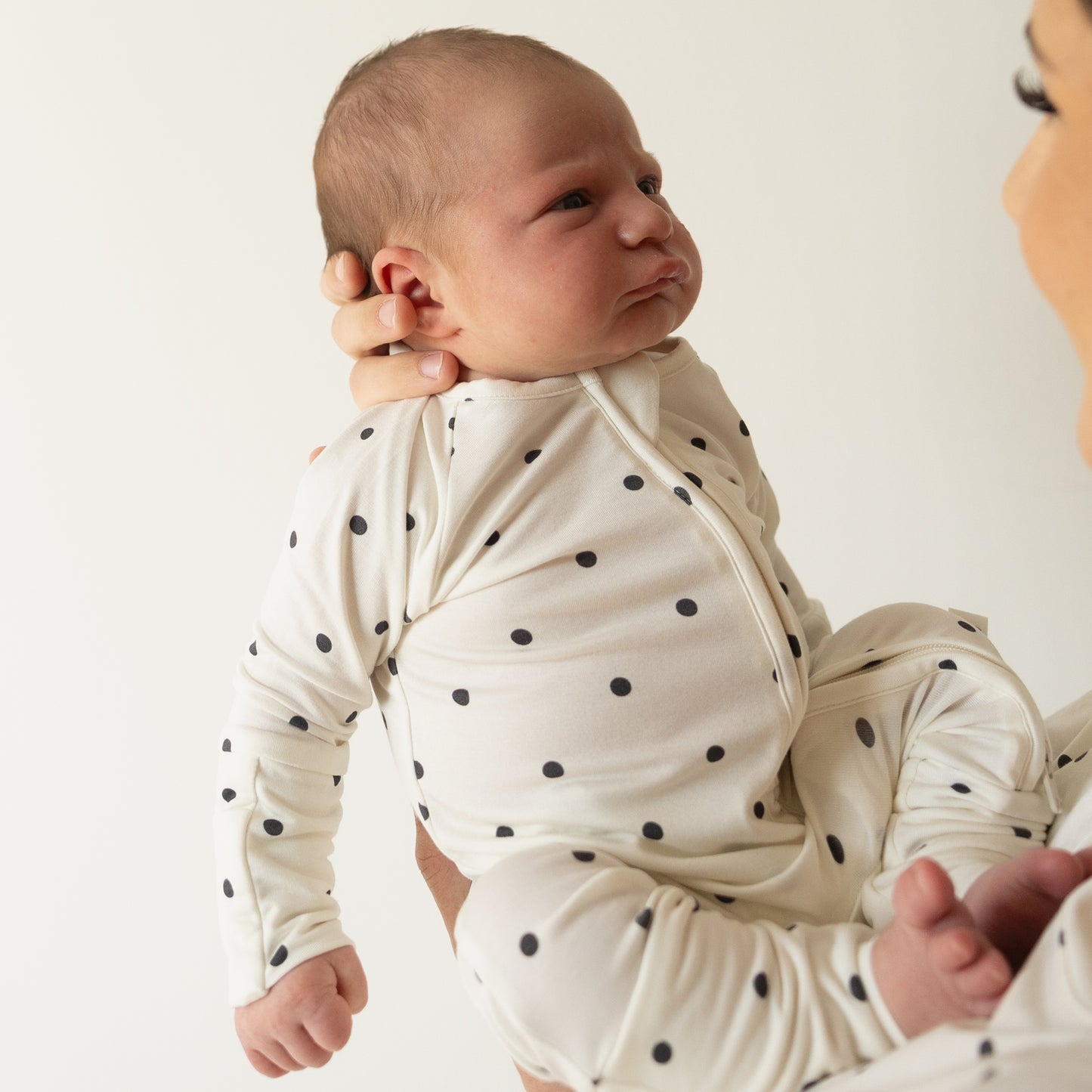 A baby in forever french baby's Bamboo Zip Pajamas | Annie—a white outfit with black polka dots—is gently held by an adult against a plain light background, the baby's cheek resting comfortably on the adult's hand.