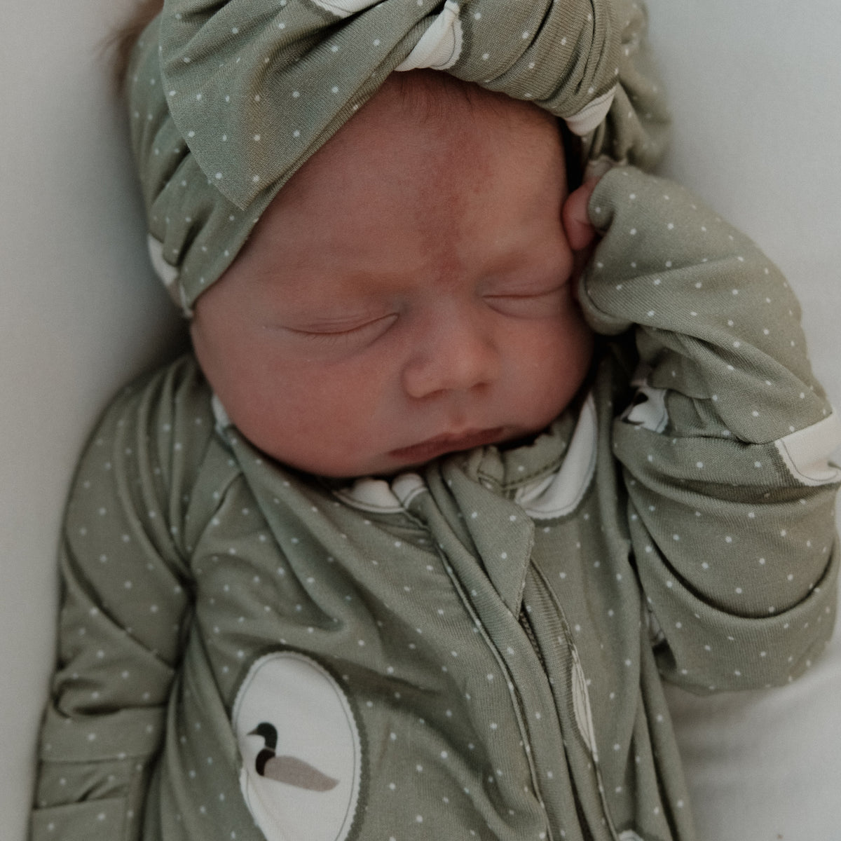 A newborn baby sleeps peacefully, dressed in cozy, hypoallergenic Bamboo Zip Pajamas | Mallard Frames by forever french baby—a green, polka-dotted outfit with bird prints and a matching headband—resting one hand near their face.