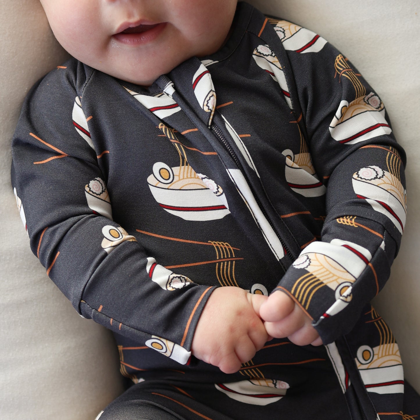 A baby in forever french baby's Bamboo Zip Pajamas | Ramen—a dark onesie with a ramen bowl print—sits clasping their hands, showing only their nose, mouth, and hands.
