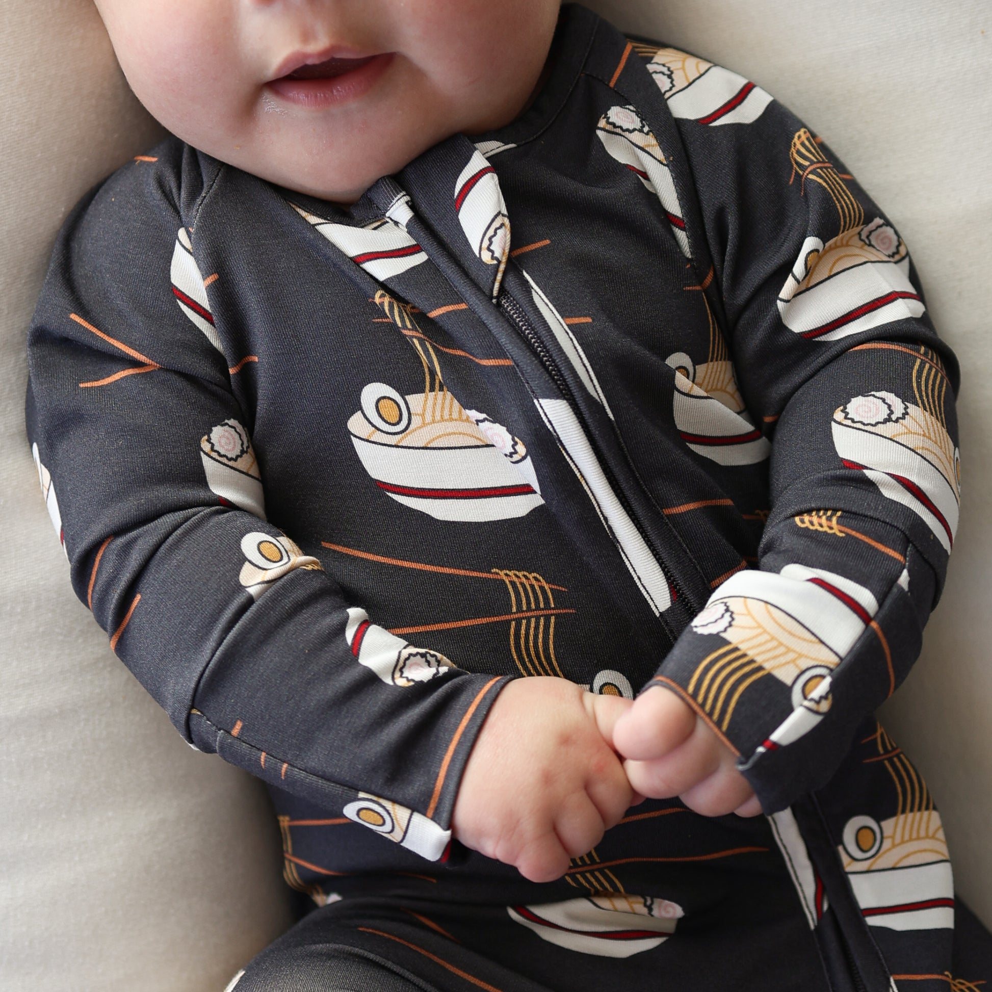 A baby in forever french baby's Bamboo Zip Pajamas | Ramen—a dark onesie with a ramen bowl print—sits clasping their hands, showing only their nose, mouth, and hands.