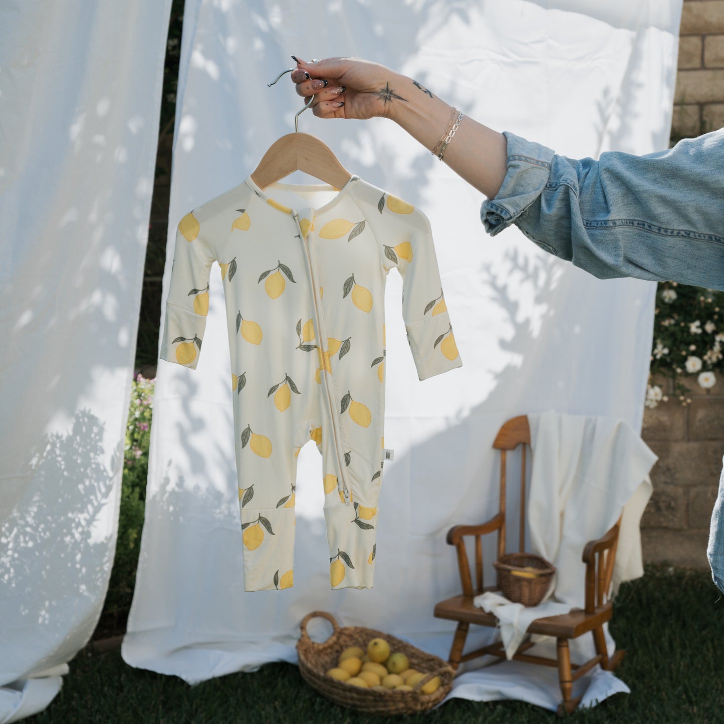A hand holds up the forever french baby Bamboo Zip Pajamas | Lemon on a wooden hanger. In the background, a rocking chair, basket of lemons, and white fabric outdoors create a perfect scene for showcasing this breathable baby sleepwear.