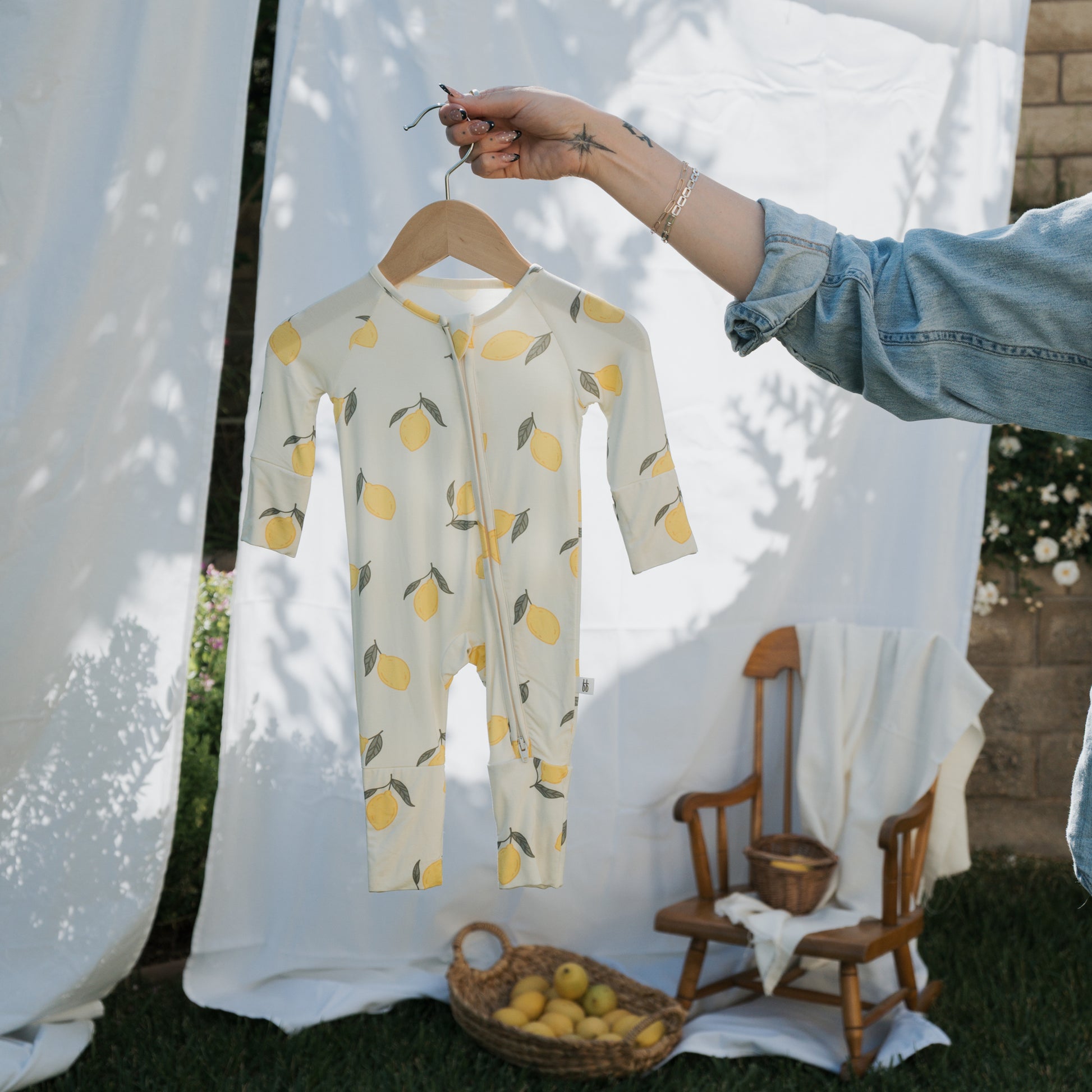 A hand holds up the forever french baby Bamboo Zip Pajamas | Lemon on a wooden hanger. In the background, a rocking chair, basket of lemons, and white fabric outdoors create a perfect scene for showcasing this breathable baby sleepwear.
