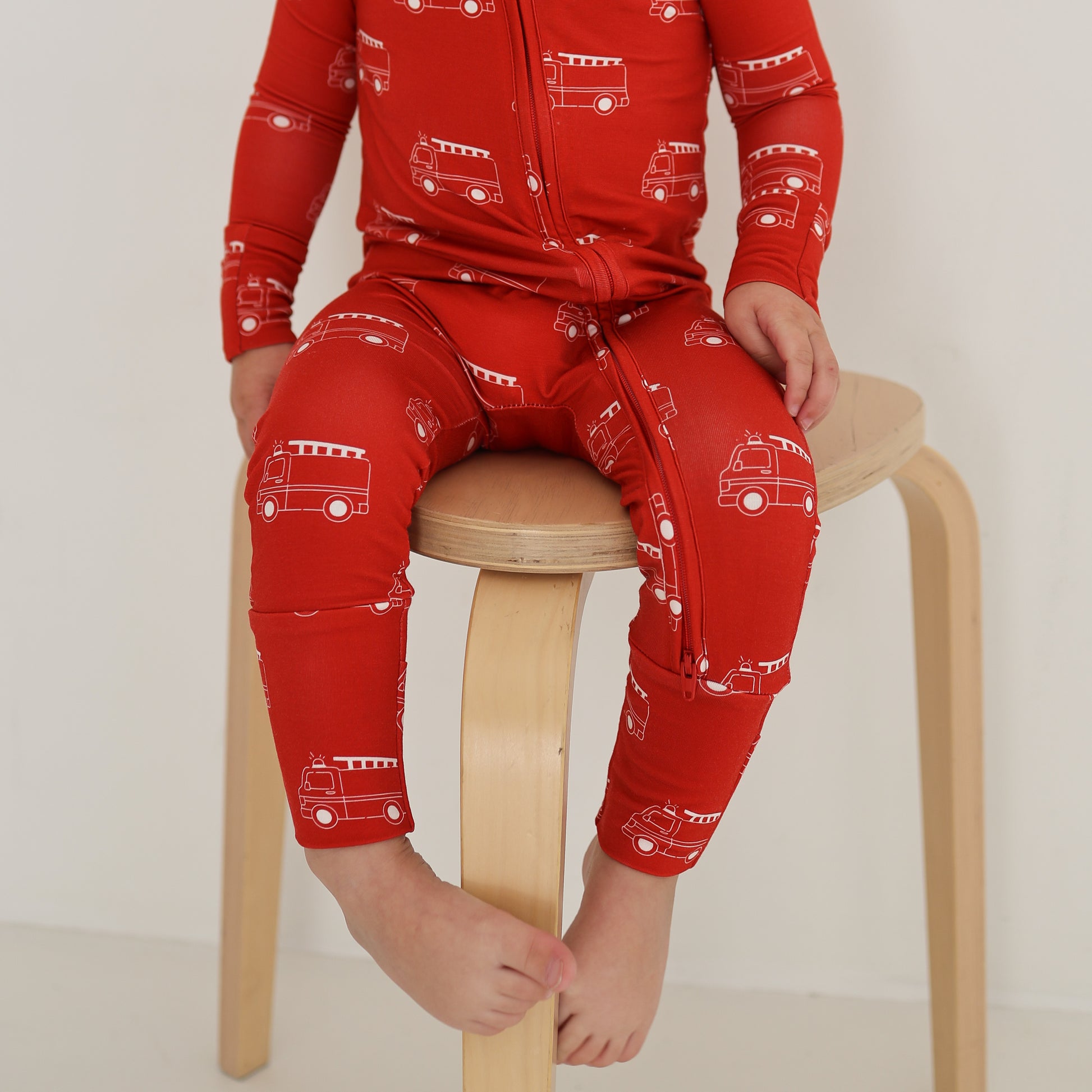 A young child models the forever french baby Bamboo Zip Pajamas | Fire Truck, sitting barefoot on a light wooden stool against a plain white background. The child's head is not visible in the image.