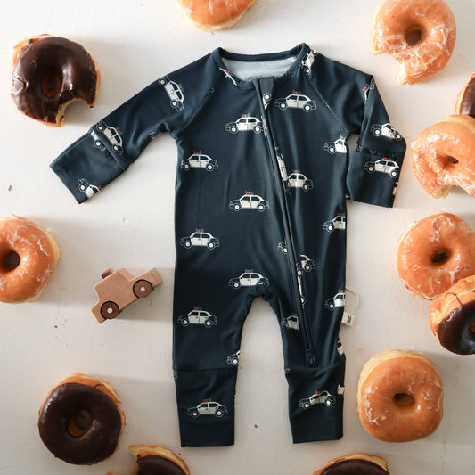 The forever french baby Bamboo Zip Pajamas | Police Cruiser, made from hypo-allergenic materials, is laid flat on a white surface surrounded by glazed and chocolate donuts and a small wooden toy car.