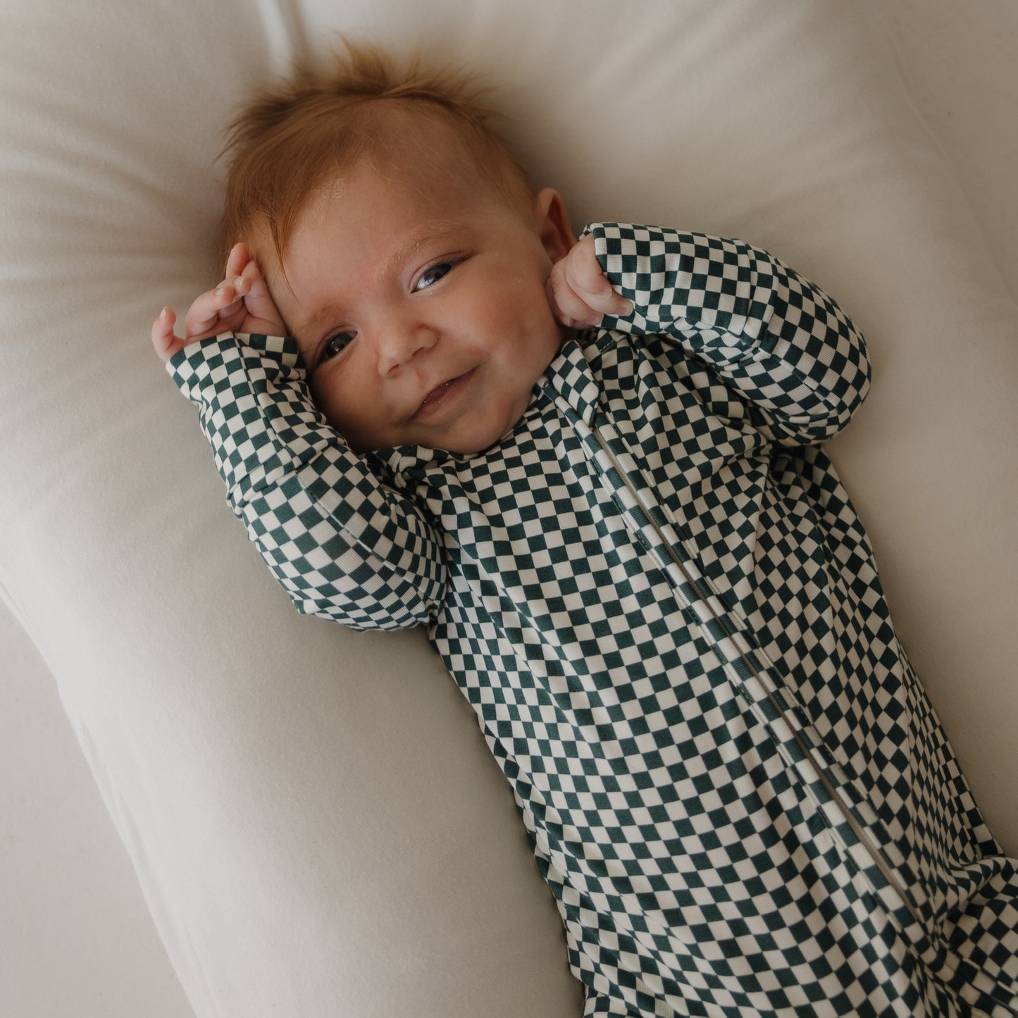A baby with light hair smiles softly on a white pillow, wearing forever french baby's Bamboo Zip Pajamas | Lucky Micro Check—a long-sleeved, breathable black-and-white checkered onesie—with hands gently raised near the head.