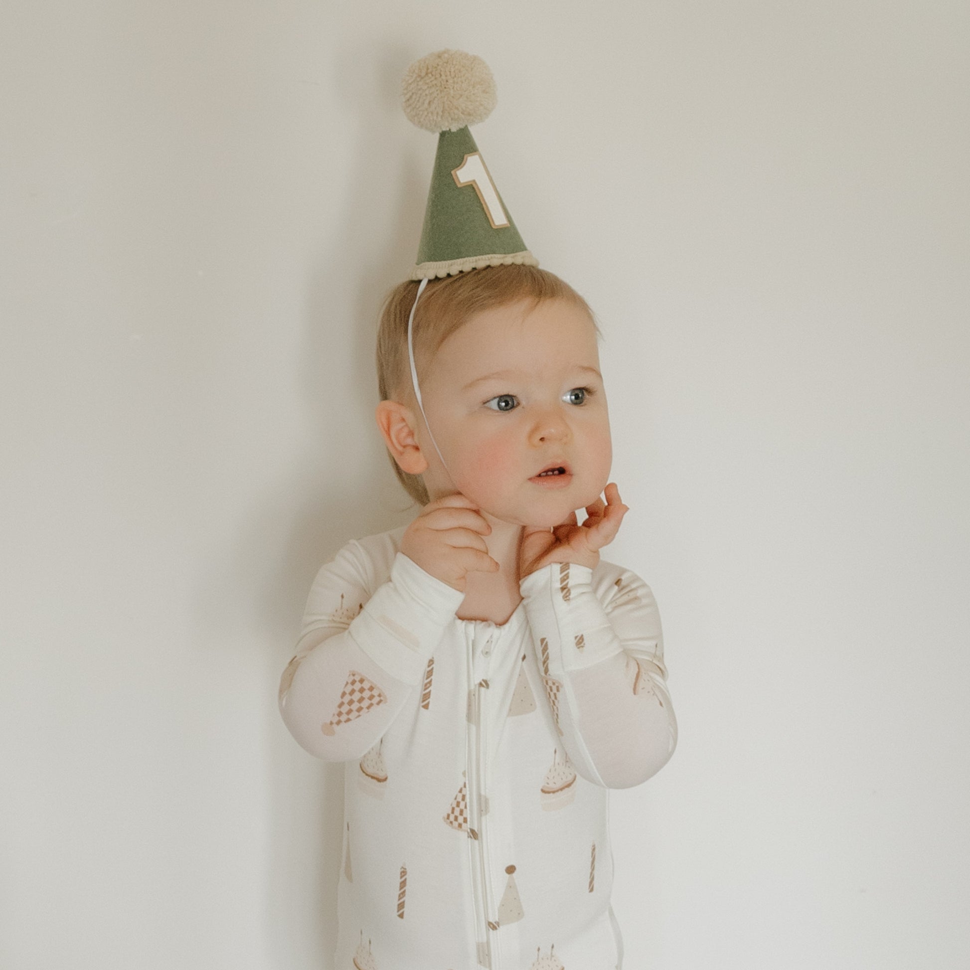 A toddler wearing forever french baby's Bamboo Zip Pajamas | Birthday Boy—a hypo-allergenic, white onesie adorned with ice cream cone prints—stands against a plain white wall, looking off to the side with hands near their face and sporting a green party hat with a “1” on it.