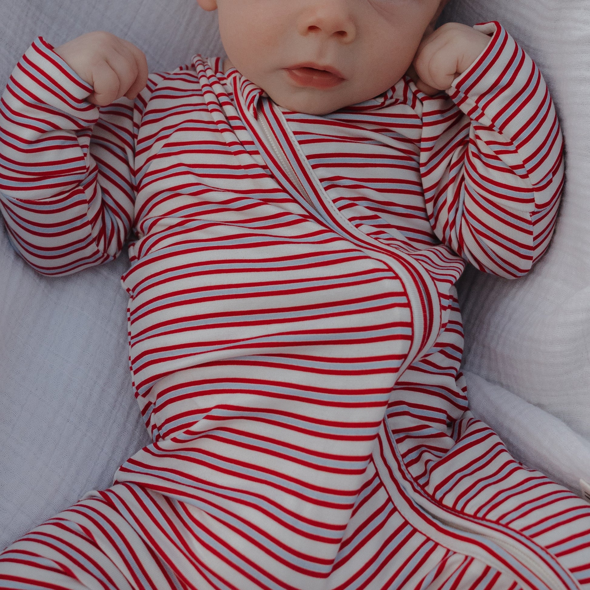 A baby wearing forever french baby's Bamboo Zip Pajamas | Heritage Stripe lies on a soft white blanket, fists slightly raised, with only the lower part of their face visible.