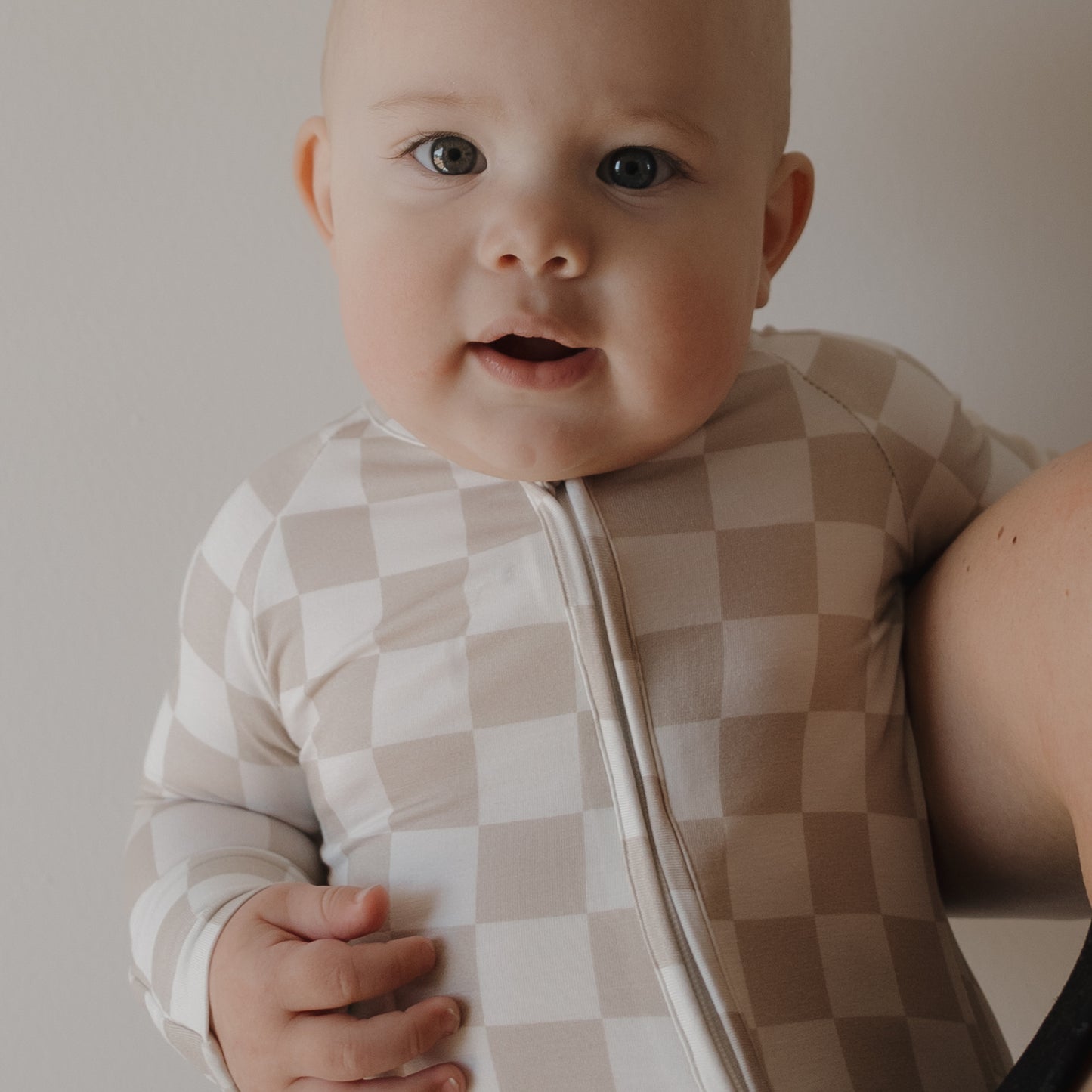 A baby in forever french baby’s Bamboo Zip Pajamas | Checkerboard, featuring a beige and white checkered pattern, gazes at the camera while resting against an adult’s arm with a neutral background.