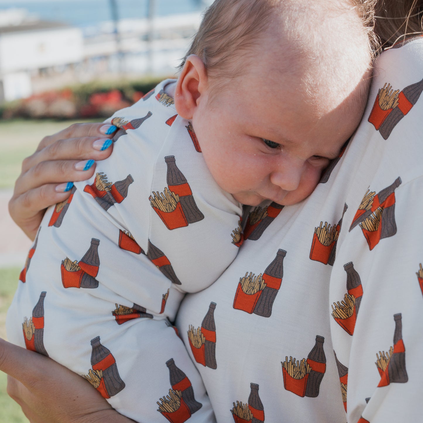 A baby and adult rest outdoors in matching forever french baby Bamboo Zip Pajamas | Fries & Cola, featuring a fries and soda bottle pattern. The adult has blue nail polish. The sunny background is slightly blurred.