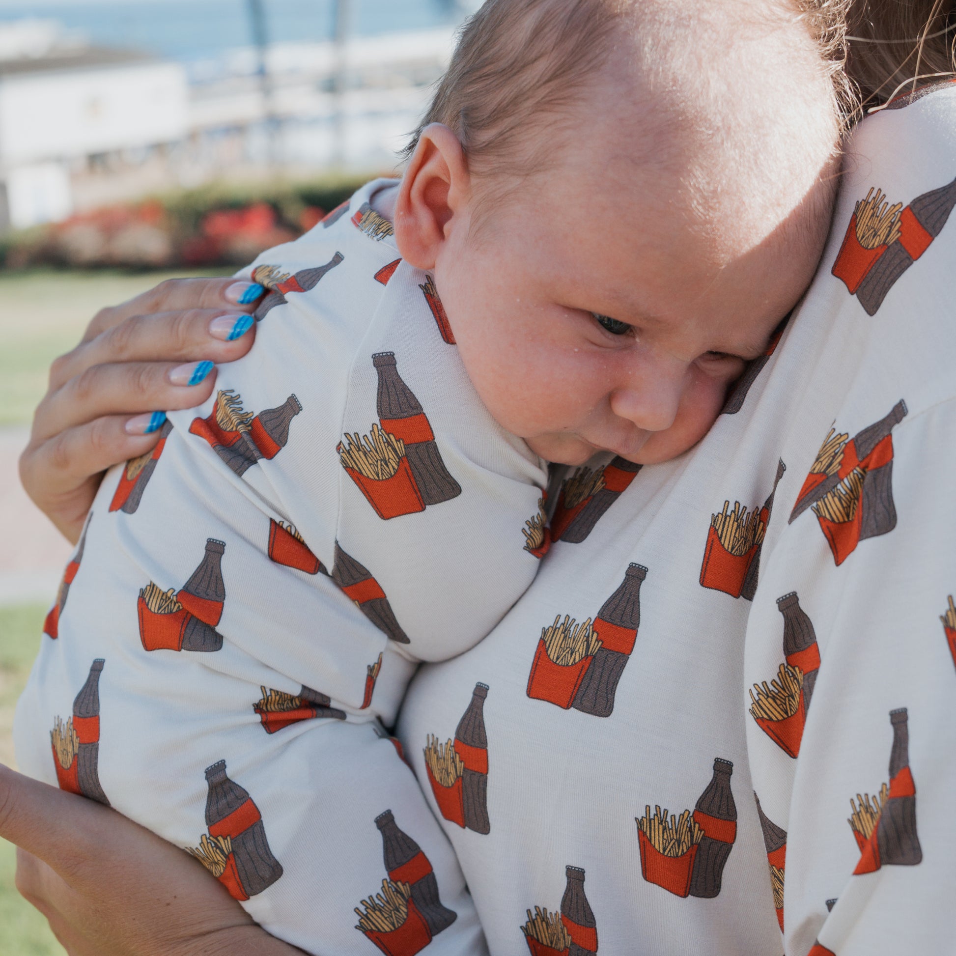 A baby and adult rest outdoors in matching forever french baby Bamboo Zip Pajamas | Fries & Cola, featuring a fries and soda bottle pattern. The adult has blue nail polish. The sunny background is slightly blurred.