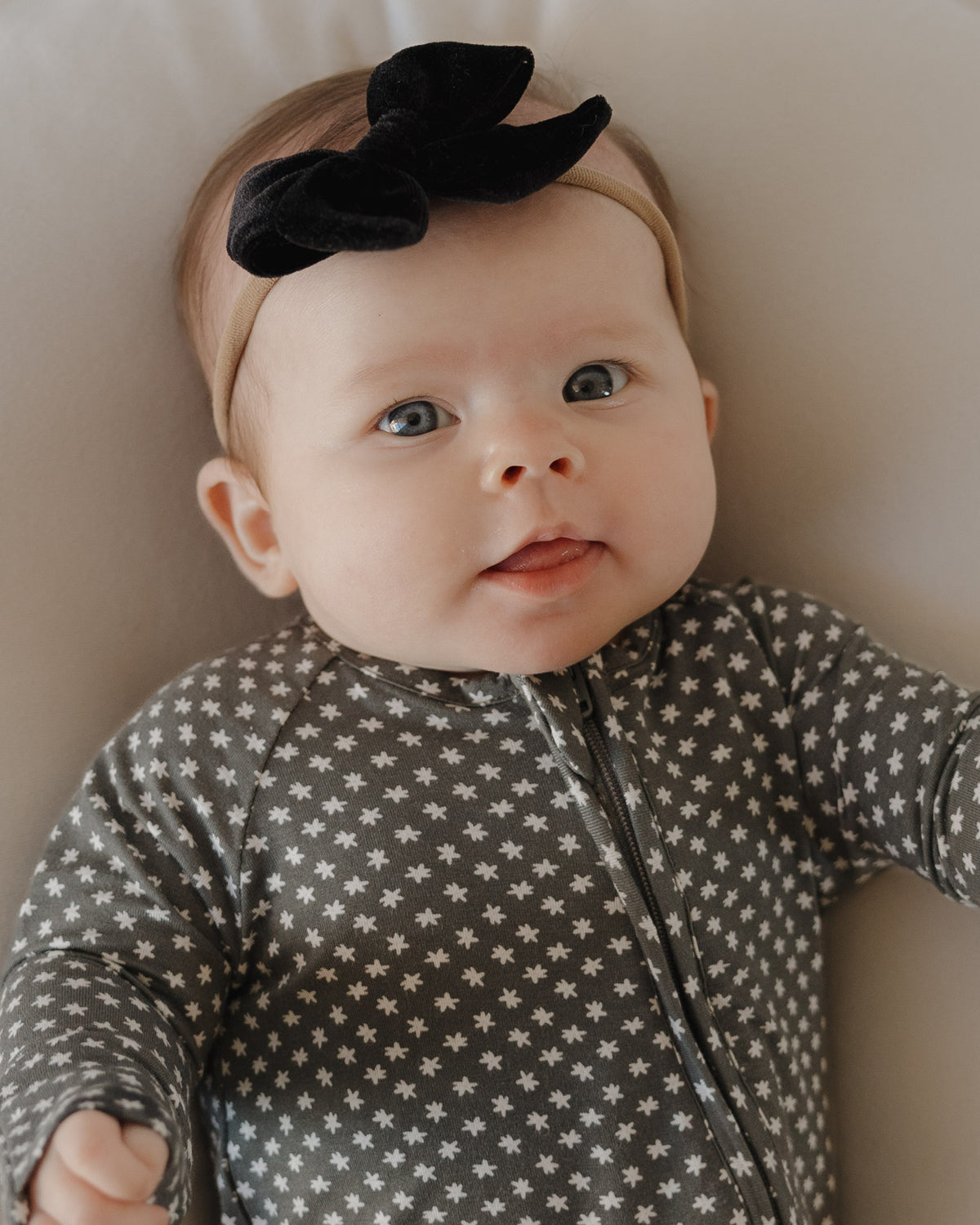 A baby lies on a soft surface, dressed in forever french baby Bamboo Zip Pajamas | Green Flowers, gazing up with wide eyes and a slight smile.