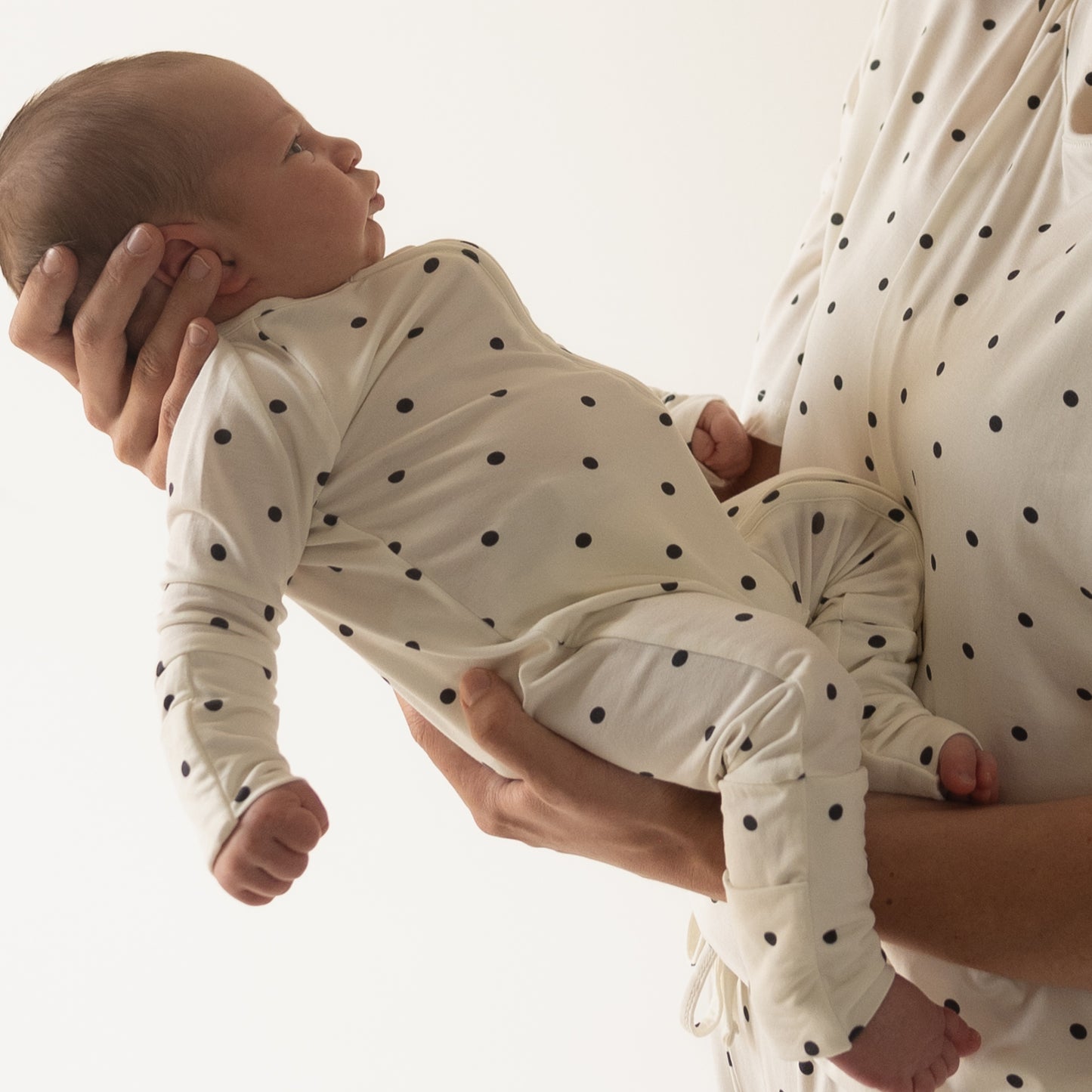 An adult holds a baby dressed in forever french baby's Bamboo Zip Pajamas | Annie. Both wear matching, breathable black polka dot bamboo outfits that stand out against a light background.