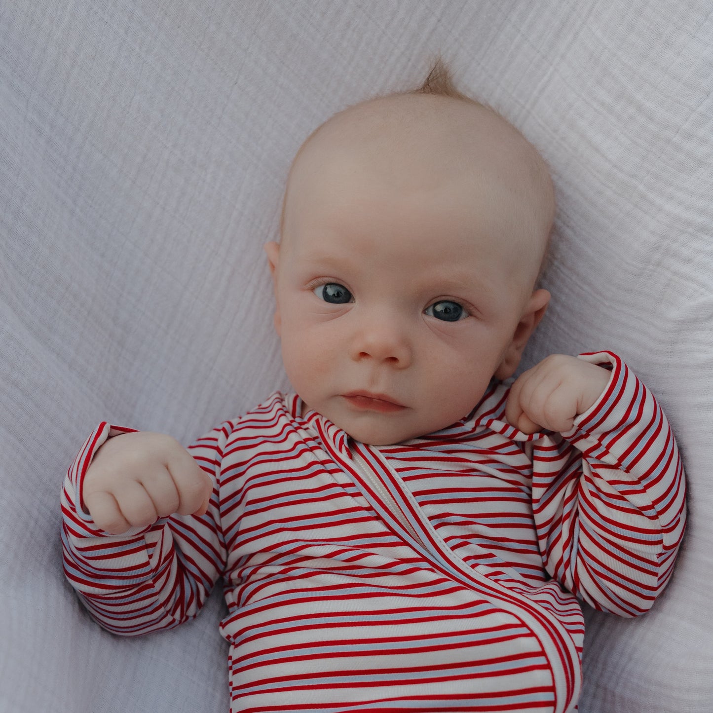 A baby in forever french baby's Bamboo Zip Pajamas | Heritage Stripe lies on a soft white blanket, looking up with wide blue eyes and slightly clenched fists, cozy in the red and white striped sleepwear.