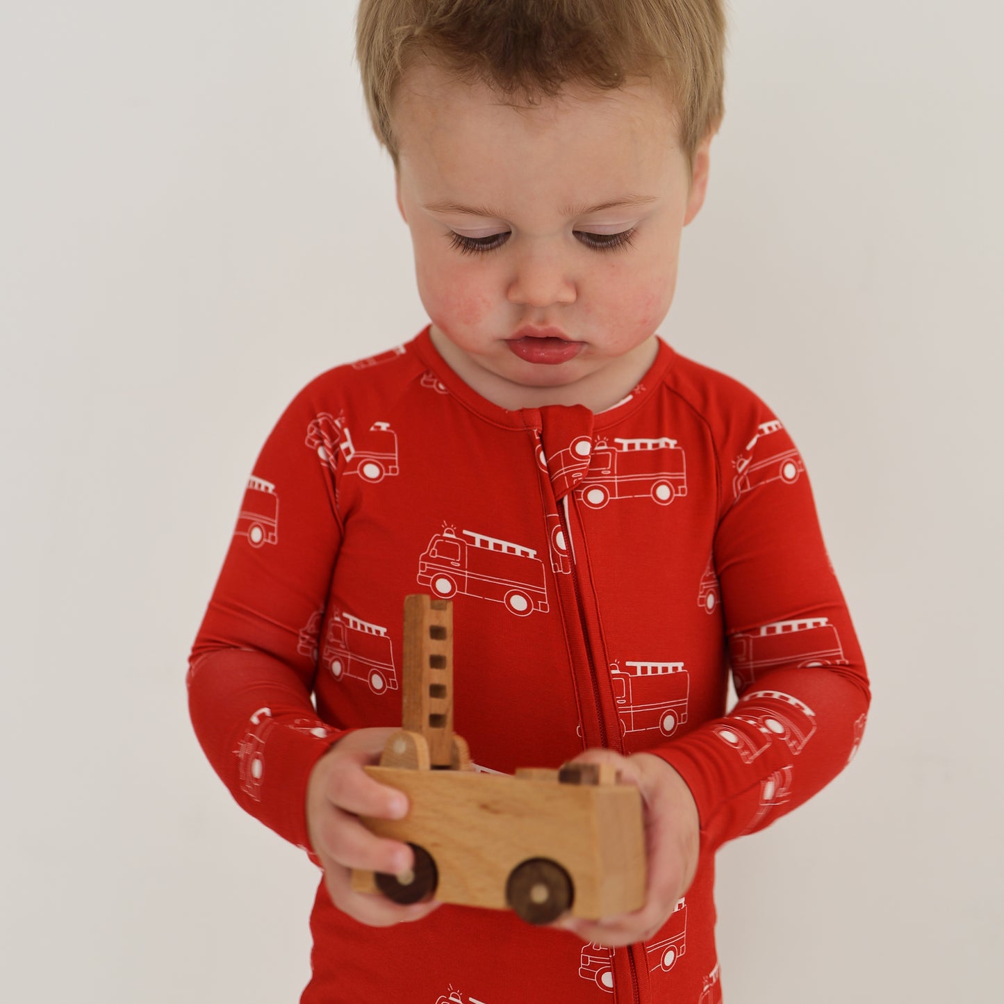 A young child in forever french baby's Bamboo Zip Pajamas | Fire Truck with fire truck prints holds a wooden toy vehicle while looking down against a plain white background.