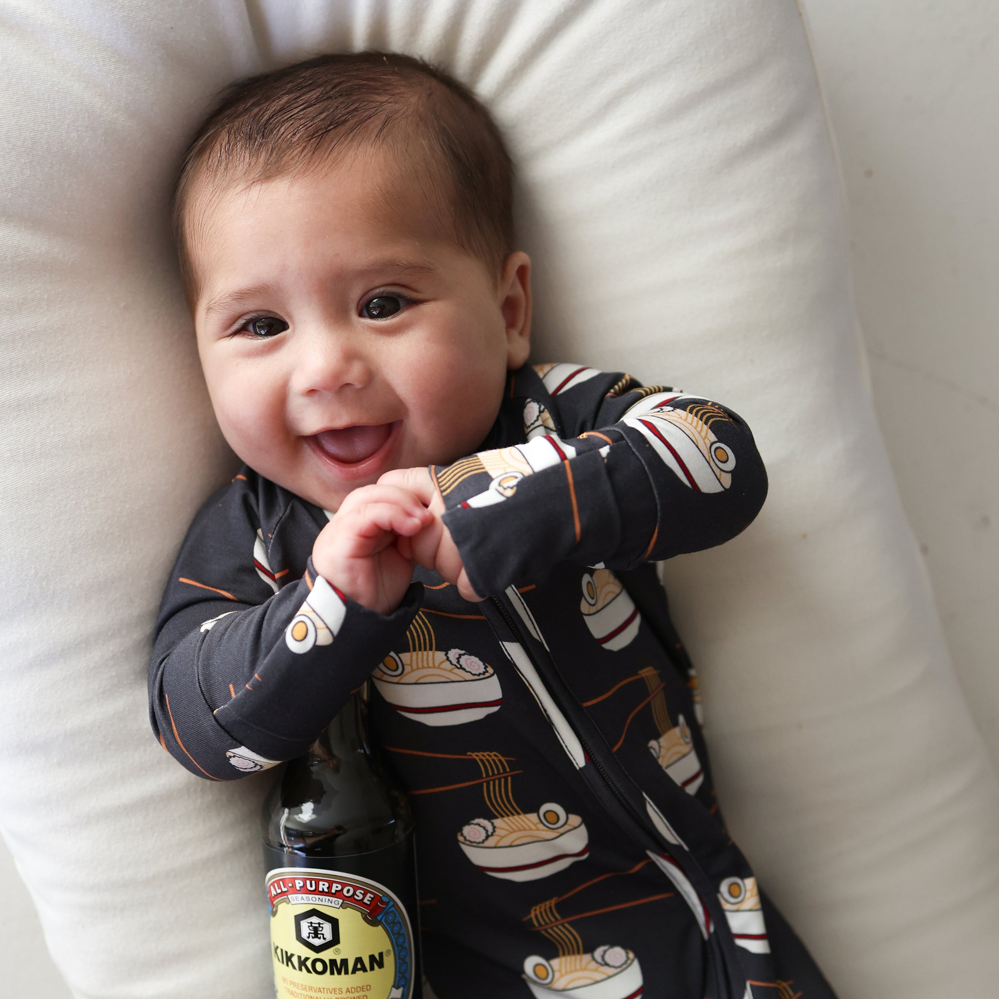 A smiling baby in forever french baby Bamboo Zip Pajamas | Ramen, featuring ramen bowl prints, lies on a white pillow while holding a bottle of Kikkoman soy sauce.