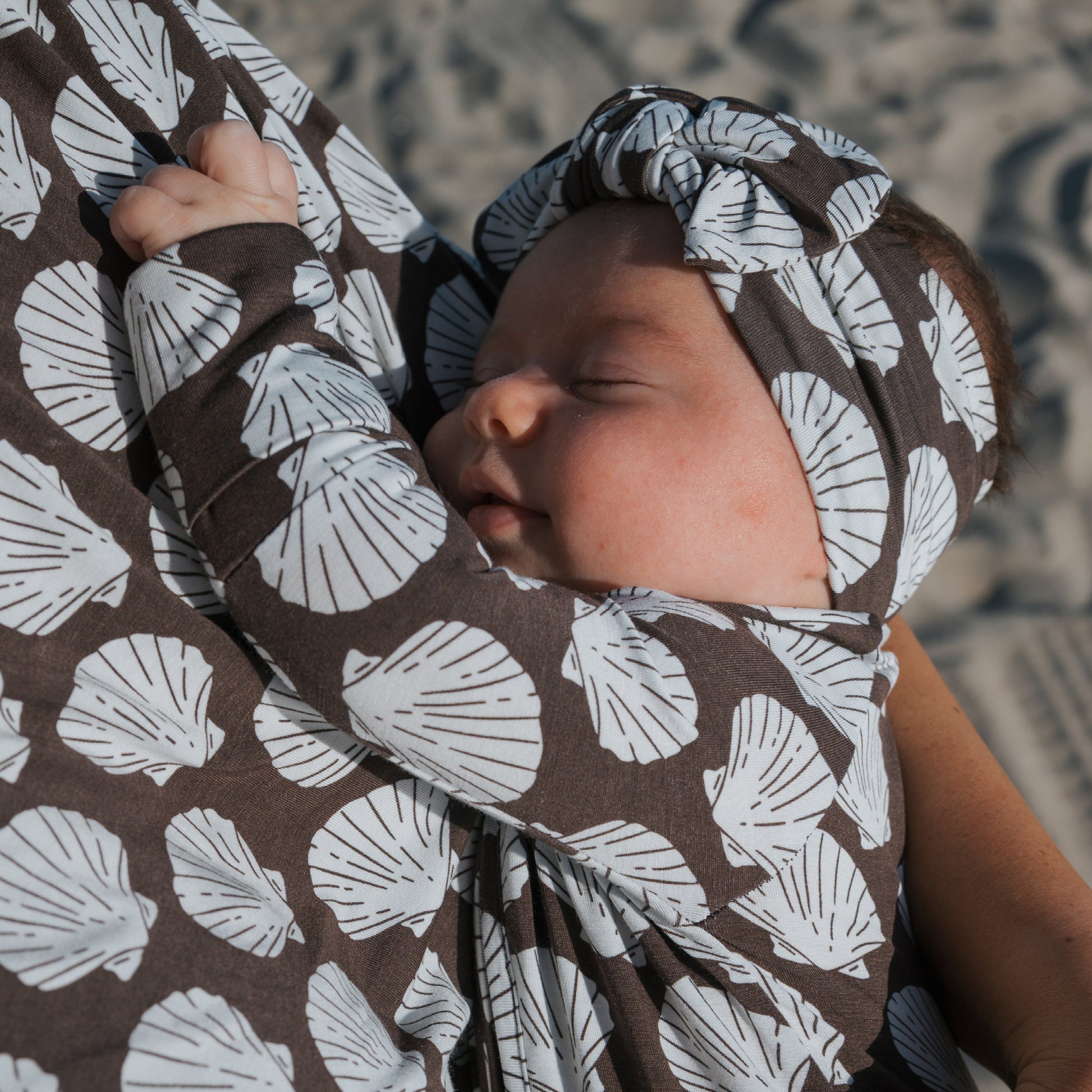 A baby sleeps peacefully in forever french baby Bamboo Zip Pajamas | Seashells, featuring a white seashell pattern and matching headband, against a sandy background.