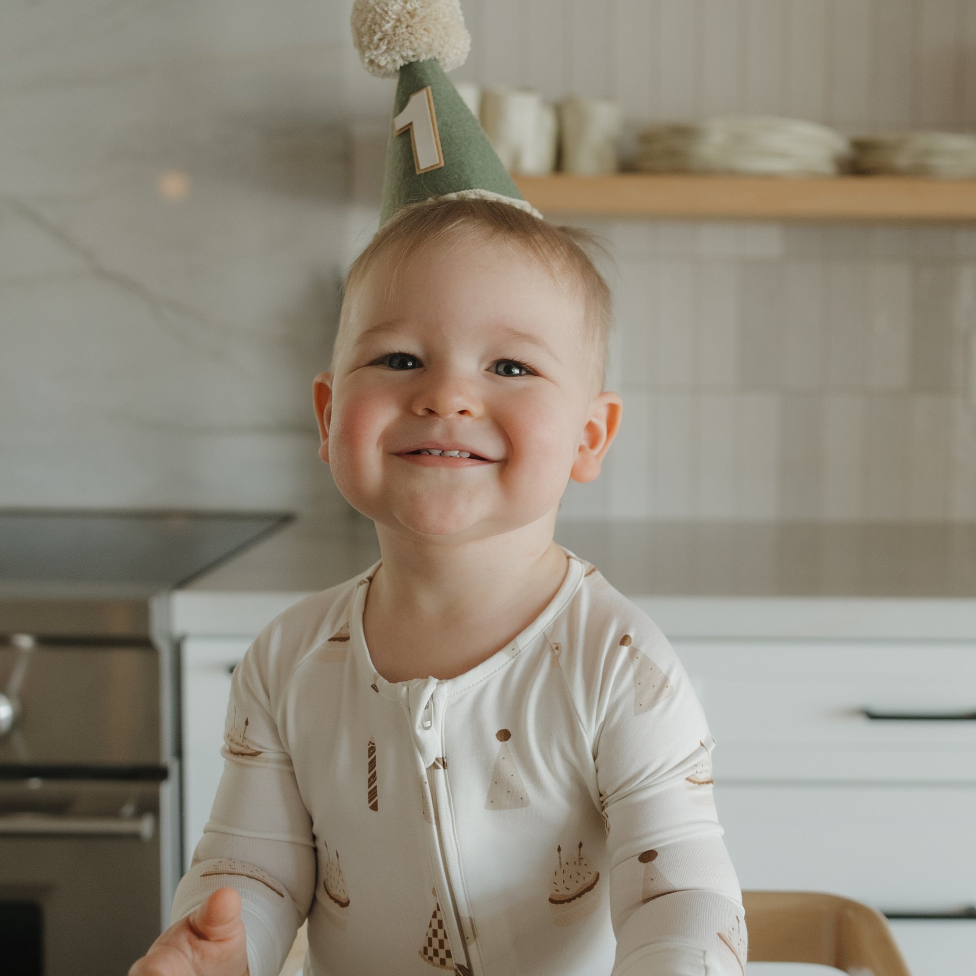 A smiling toddler wearing a birthday hat with the number one on it stands in the kitchen dressed in forever french baby’s Bamboo Zip Pajamas | Birthday Boy—a soft and breathable white onesie with festive birthday cake prints that’s as comfy as it is adorable.