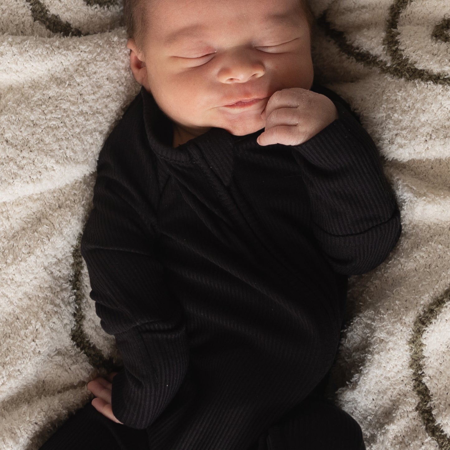 A newborn wearing forever french baby's Bamboo Zip Pajamas in Ribbed Black lies on a soft, swirl-patterned blanket, eyes closed and one hand gently touching their face.