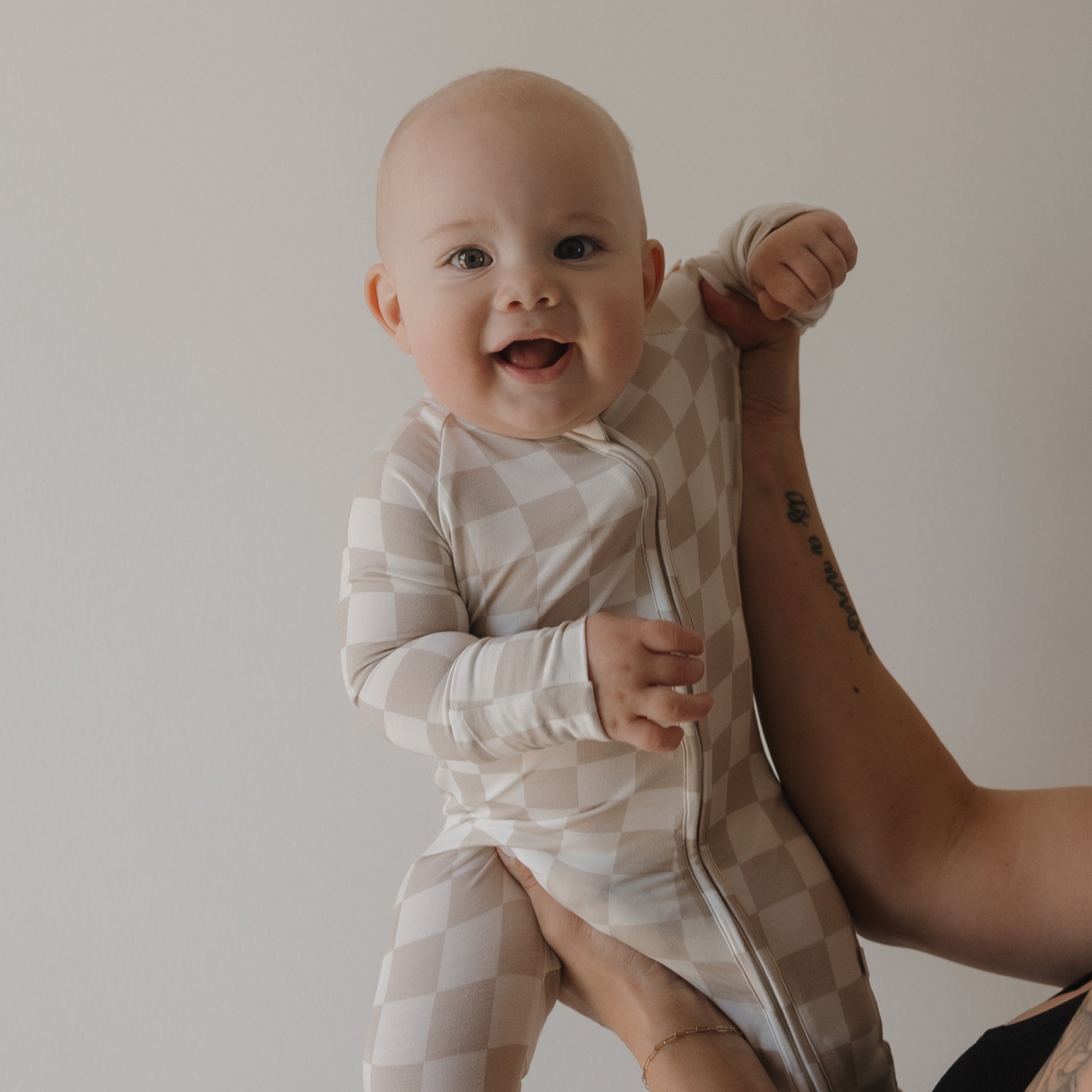 A smiling baby wears the forever french baby Bamboo Zip Pajamas | Checkerboard in beige and white, held by an adult with a tattooed arm. The hypoallergenic onesie stands out against a plain, light background.