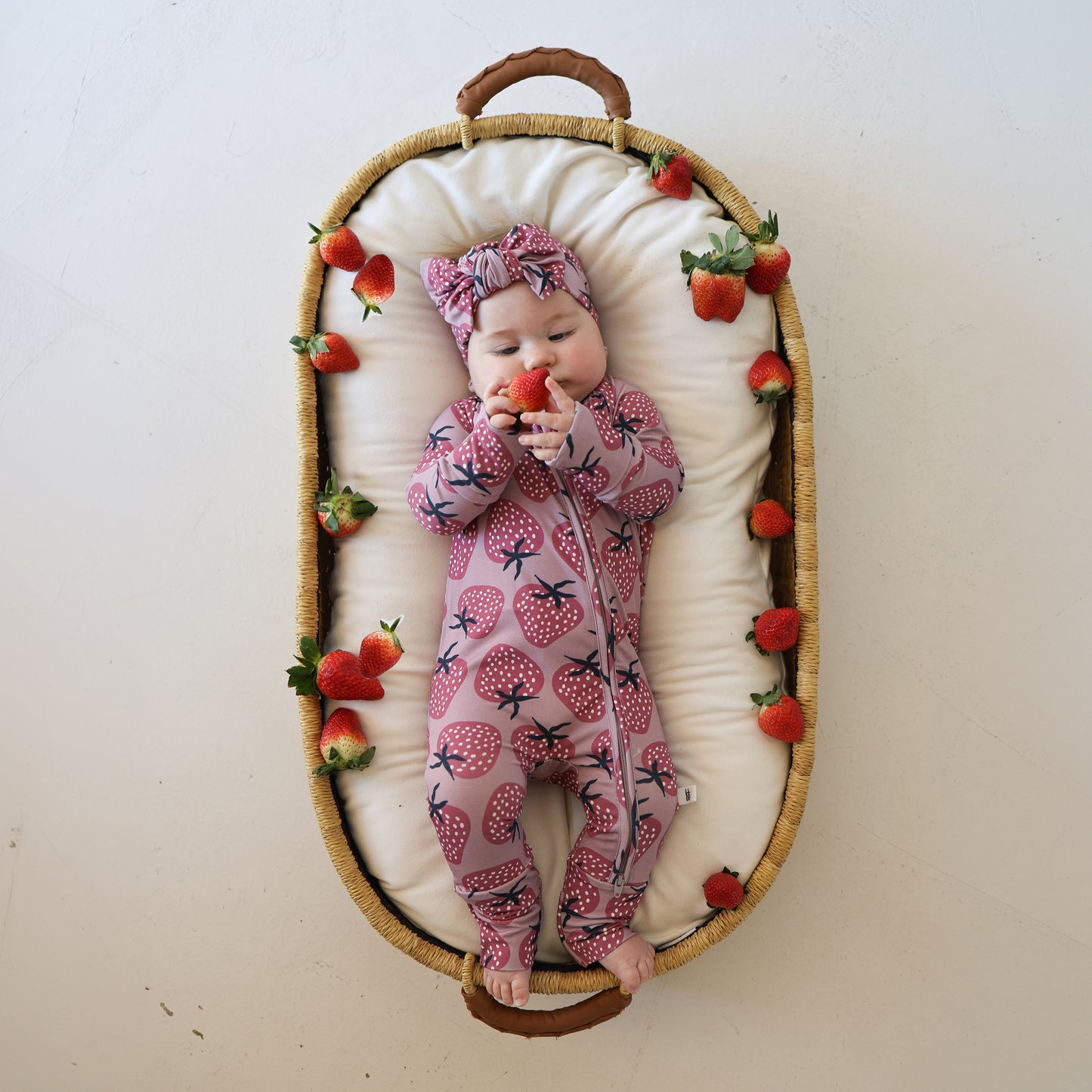 A baby in forever french baby’s Bamboo Zip Pajamas | Matilda—a pink strawberry-patterned onesie with a matching headband—lies in a cushioned basket among fresh strawberries, holding one up with both hands.