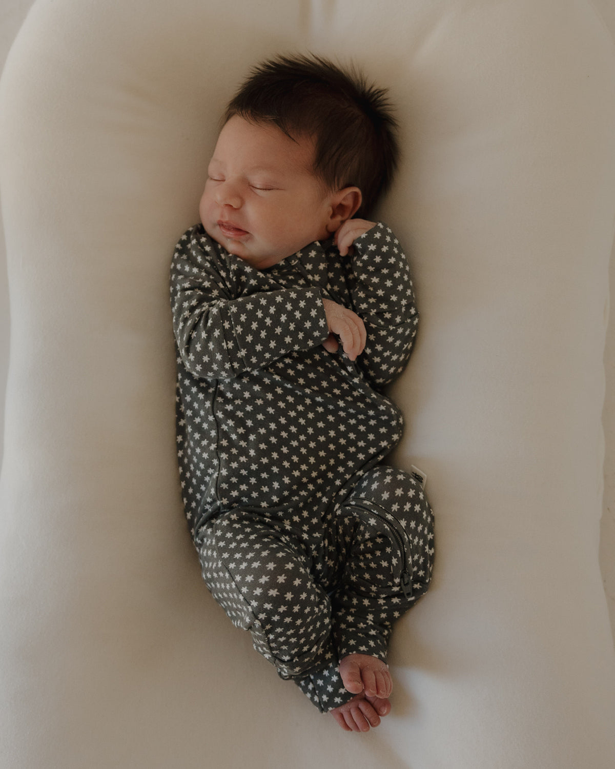 A newborn with dark hair sleeps peacefully on a white cushion, wearing forever french baby's Bamboo Zip Pajamas in the Green Flowers print.