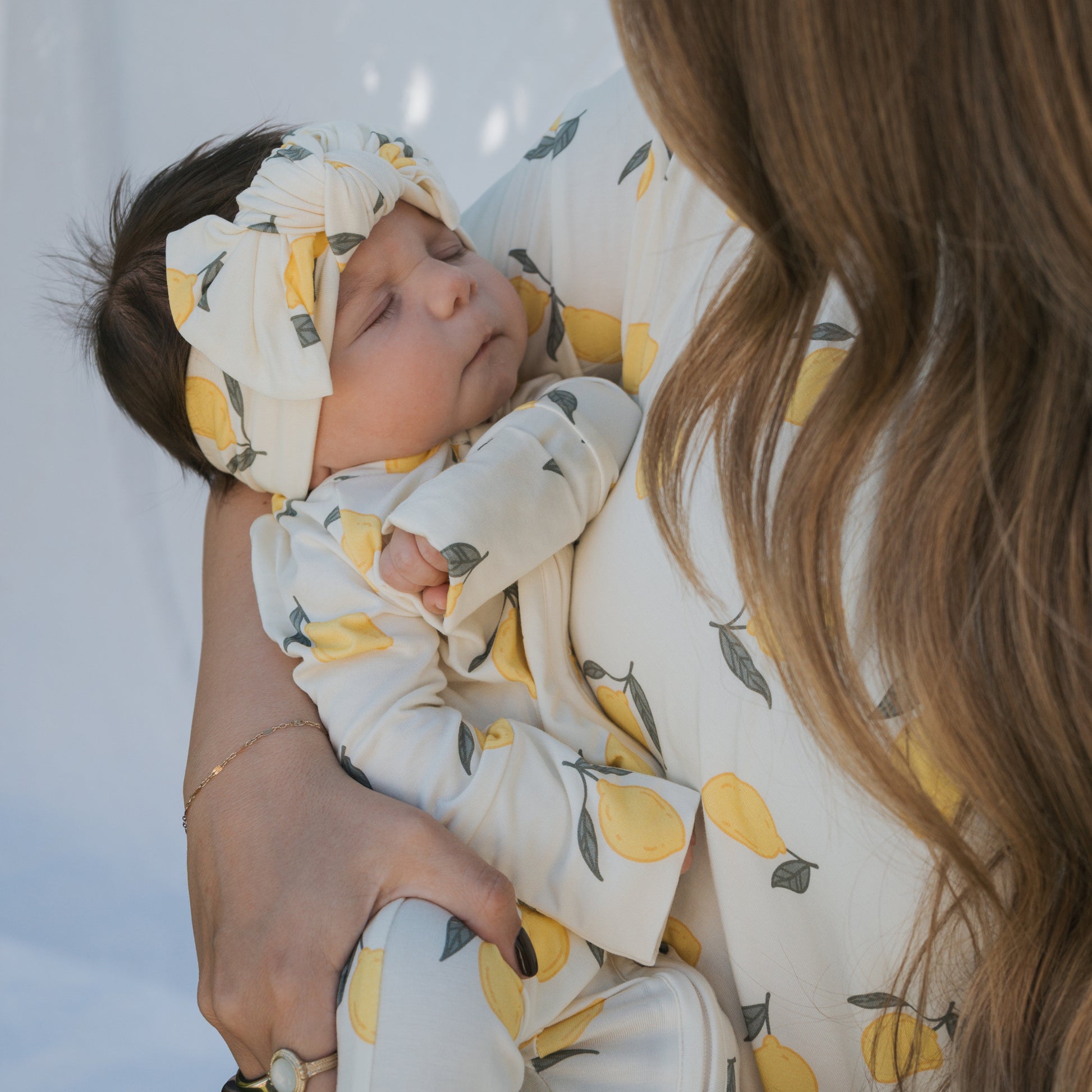 A woman with long brown hair cradles a sleeping baby. Both wear forever french baby’s Bamboo Zip Pajamas | Lemon, featuring soft, breathable bamboo fabric and a matching lemon print for cozy and comfortable sleepwear.