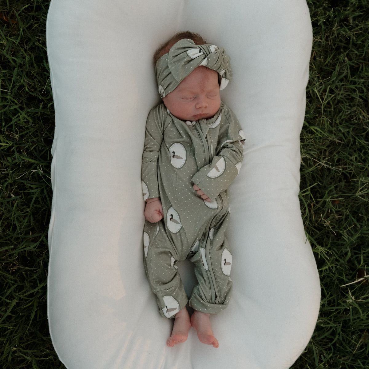 A baby dressed in forever french baby’s Bamboo Zip Pajamas in the Mallard Frames print, complete with a matching headband, sleeps peacefully on a white cushion placed on grass.