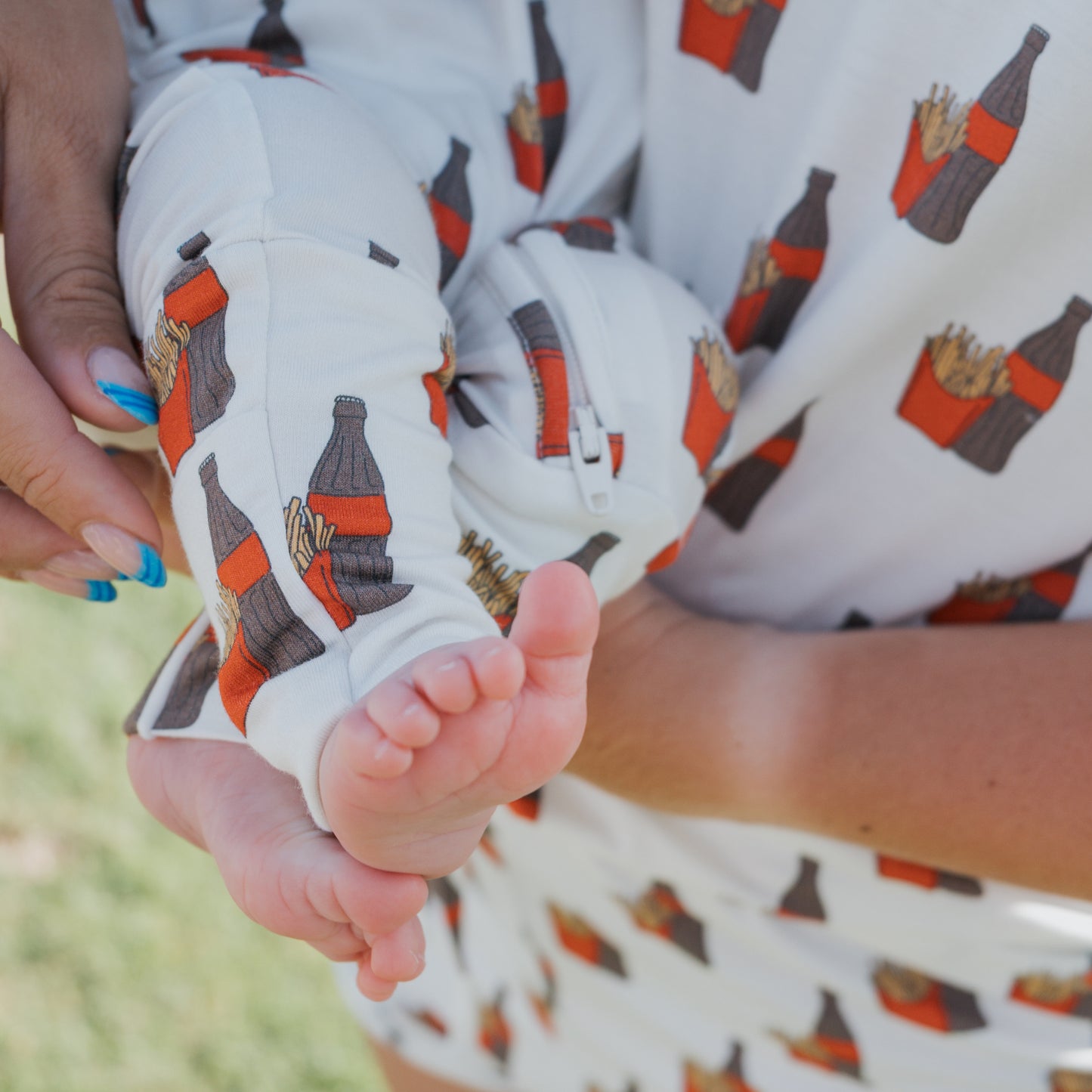 Close-up of an adult holding a baby, both in forever french baby Bamboo Zip Pajamas | Fries & Cola. The baby's bare feet and legs are visible, emphasizing the cozy, breathable design with playful soda bottle and French fry prints.