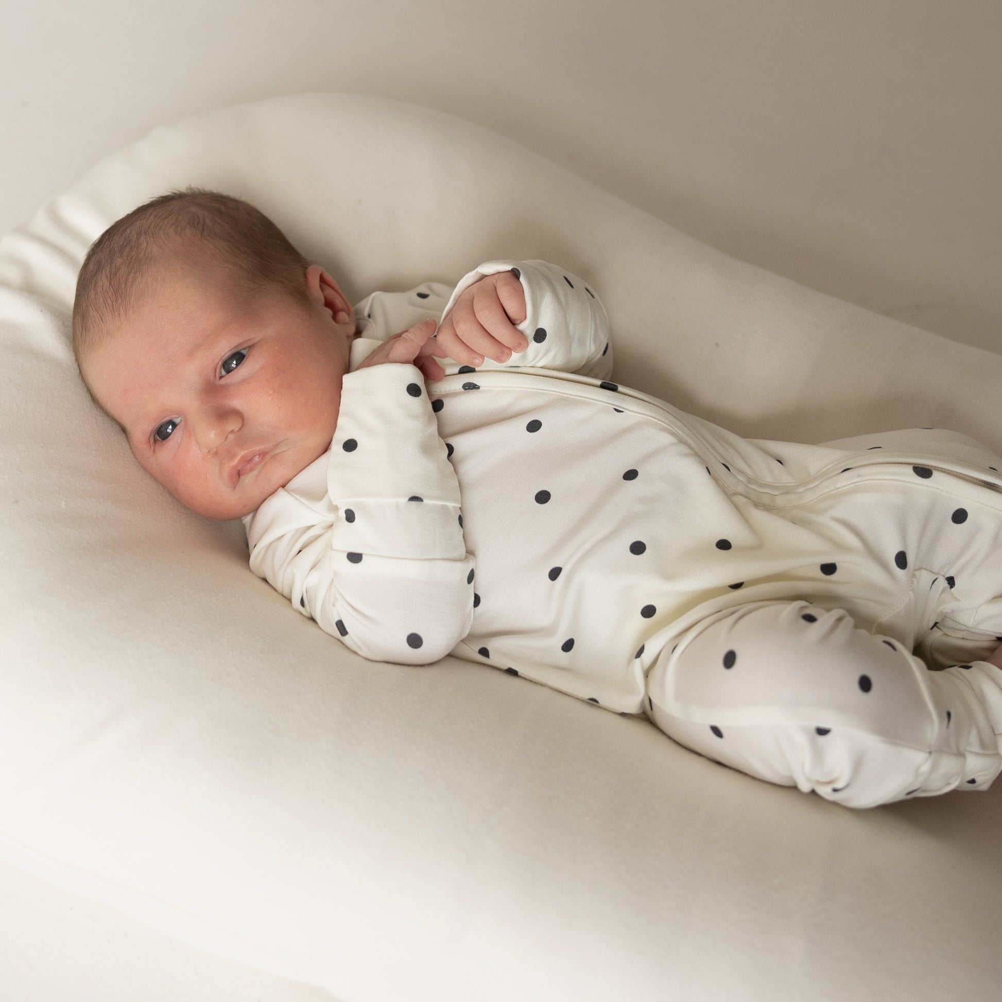 A baby wearing forever french baby Bamboo Zip Pajamas | Annie—a white onesie with black polka dots—rests on a soft, white cushion, looking up and moving its hands gently.