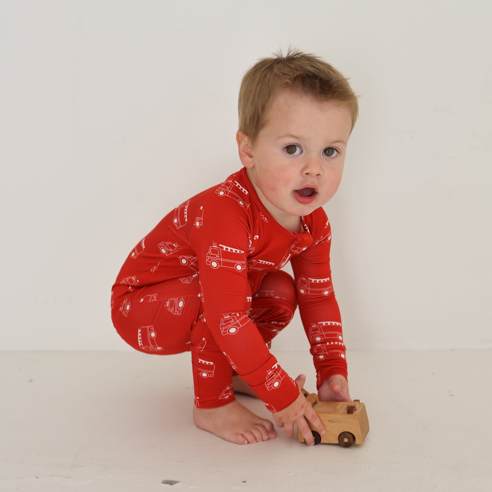 A young child wears forever french baby Bamboo Zip Pajamas | Fire Truck—red, with white vehicle motifs—while playing with a small wooden toy car against a plain white background.