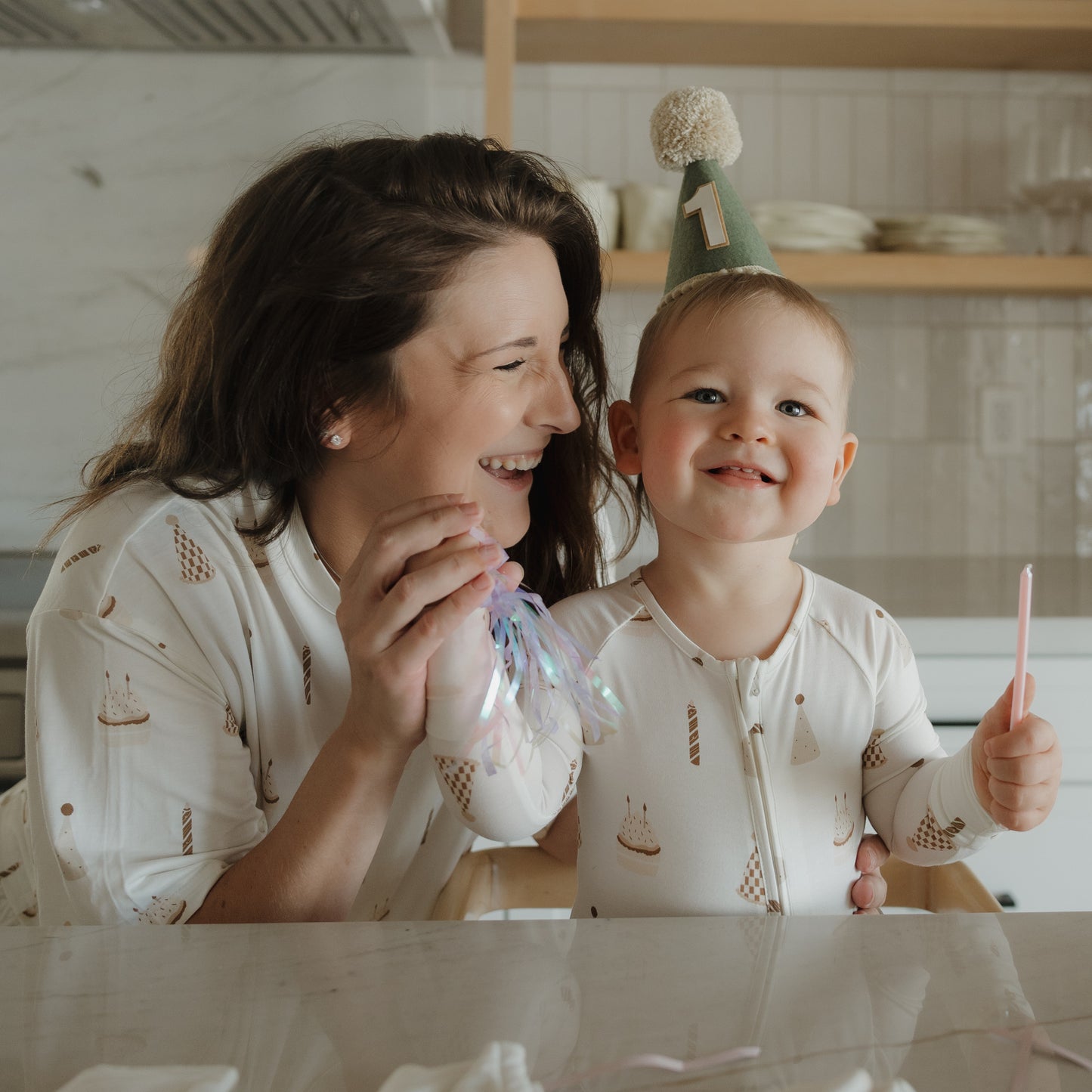 A woman and a smiling toddler wearing a "1" birthday hat sit together in the kitchen, both dressed in matching forever french baby Bamboo Zip Pajamas | Birthday Boy. The toddler holds a pink spoon and the woman holds a shiny party favor, their cheerful celebration made even cozier by the soft, breathable bamboo sleepwear.