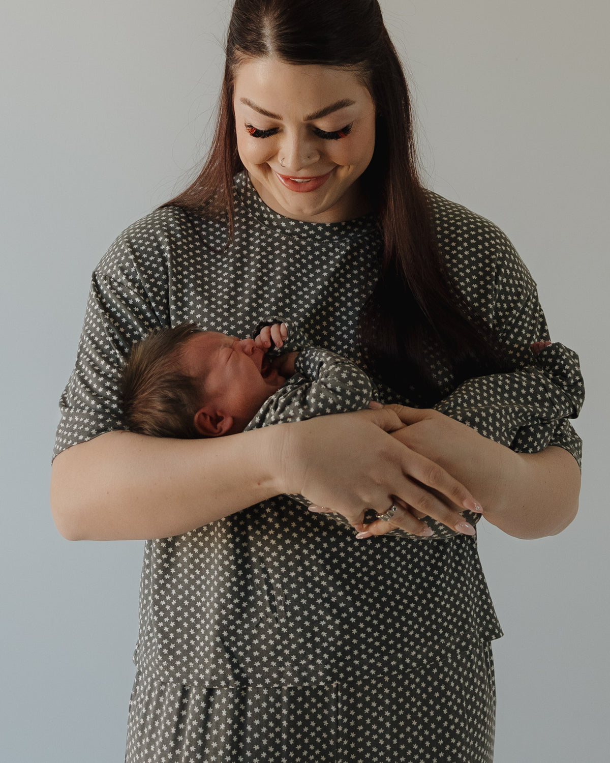 A woman with long dark hair smiles at a newborn in her arms. Both wear forever french baby's Bamboo Zip Pajamas | Green Flowers, featuring a matching hypoallergenic design with white polka dots, set against a plain light background.