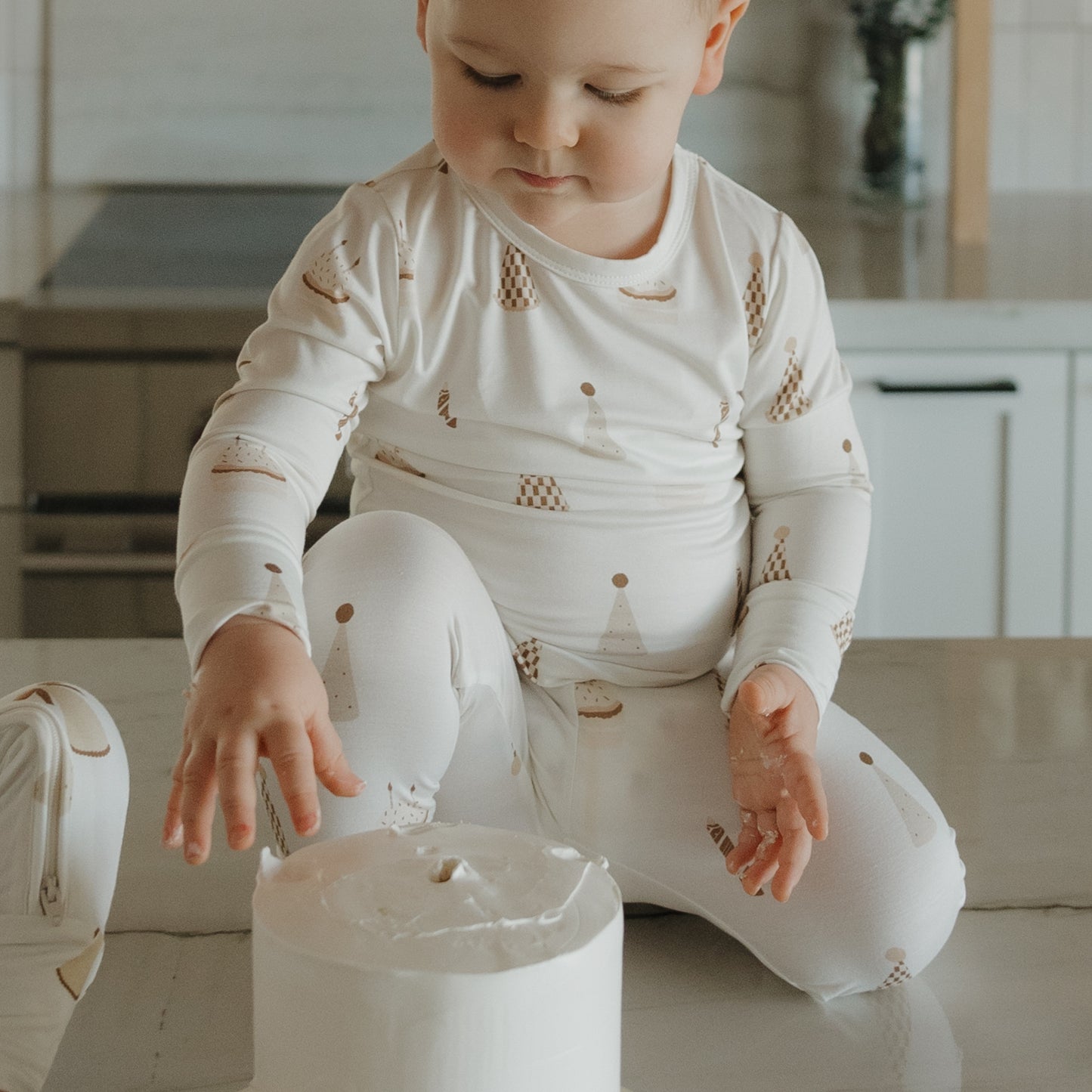 Wearing the forever french baby Child Sleepy Time Set | Birthday Boy, a toddler sits on a kitchen counter and reaches out to touch a large roll of toilet paper placed in front of them.