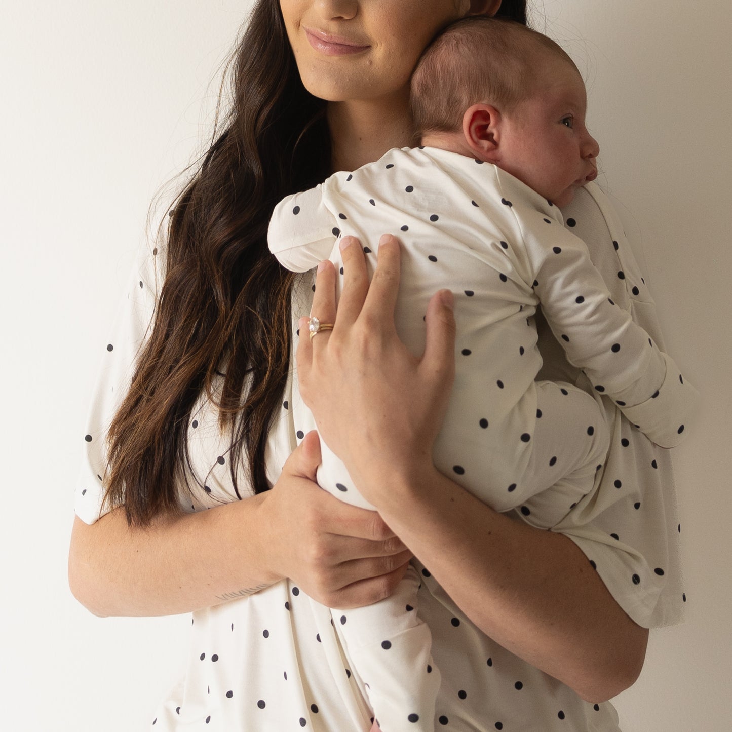A woman with long brown hair holds a baby against her chest. Both wear matching forever french baby Women's Bamboo Short Set | Annie, featuring oversized t-shirts and shorts with black polka dots, posed before a plain white background.