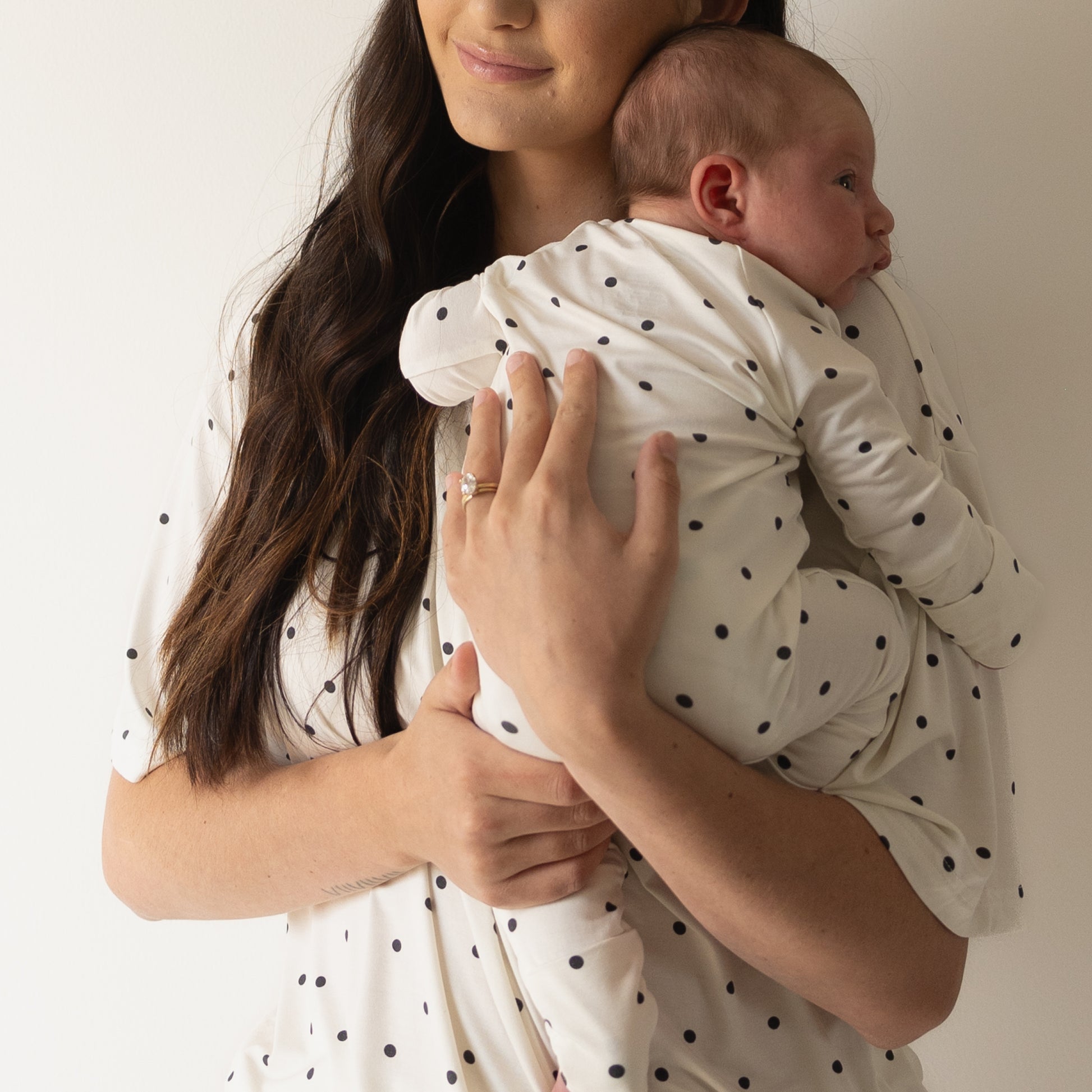 A woman with long brown hair holds a baby against her chest. Both wear matching forever french baby Women's Bamboo Short Set | Annie, featuring oversized t-shirts and shorts with black polka dots, posed before a plain white background.