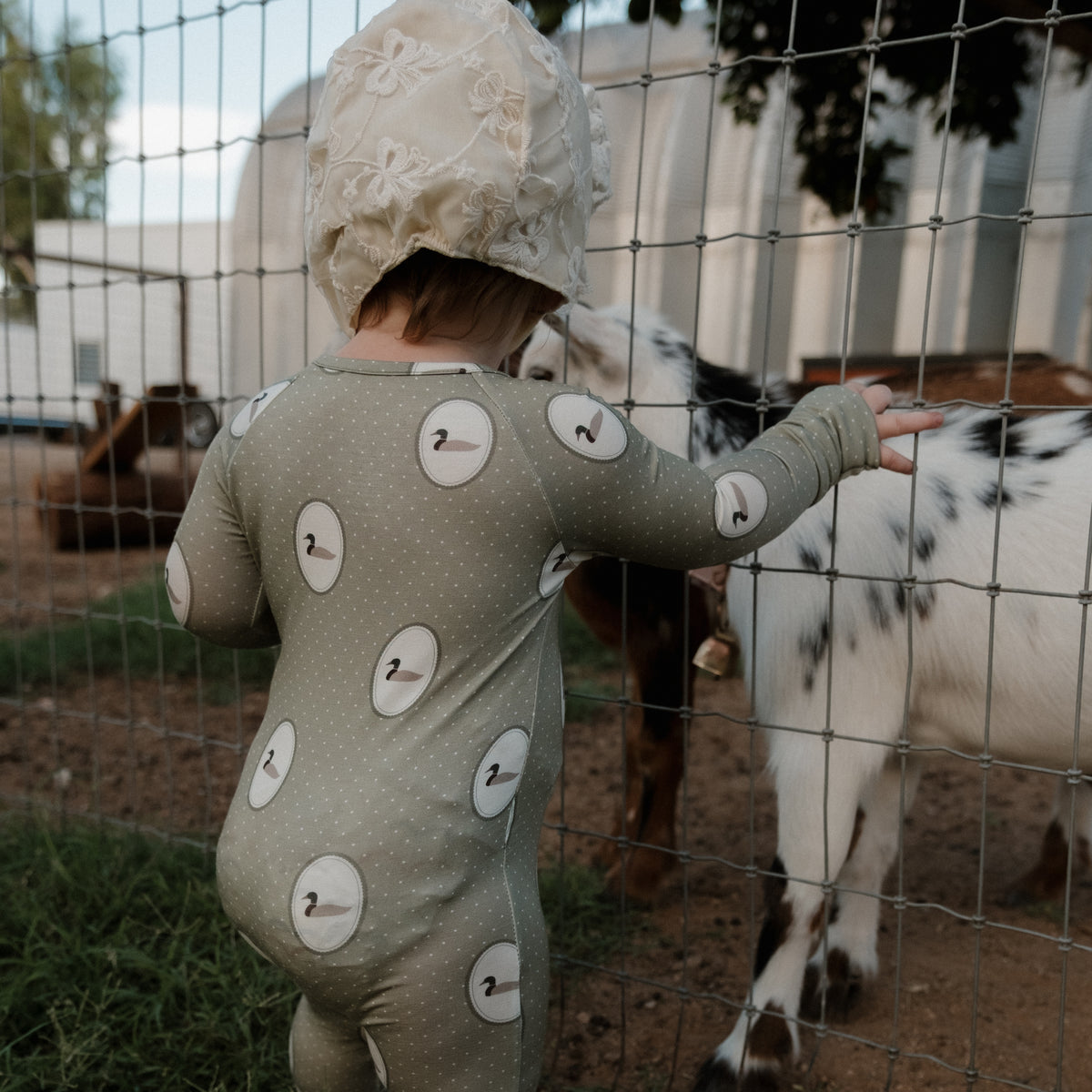 A toddler dressed in forever french baby’s Bamboo Zip Pajamas | Mallard Frames, a soft, hypoallergenic green onesie featuring a swan pattern and matching bonnet, stands by a wire fence and reaches out to touch a white and brown goat on the other side.