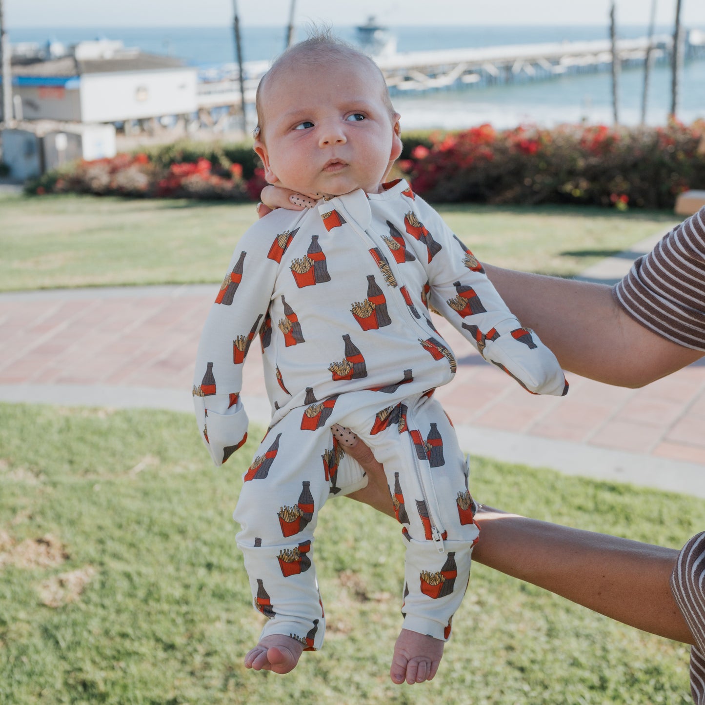 An adult holds up a baby outdoors in forever french baby's Bamboo Zip Pajamas | Fries & Cola, featuring breathable fabric and cute red fries prints, with grass, flowers, and a seaside pier in the picturesque background.