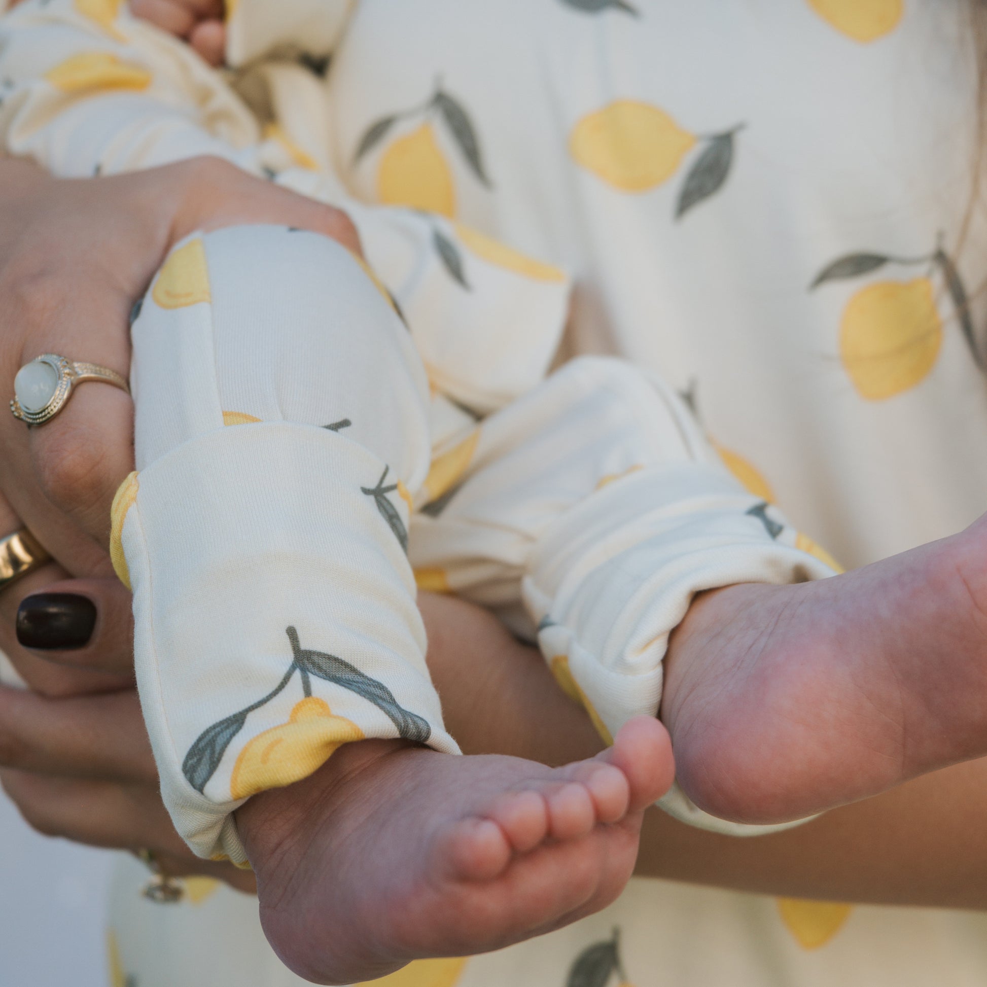 A person with painted nails and rings gently holds a baby wearing forever french baby’s Bamboo Zip Pajamas | Lemon. The hypoallergenic, breathable pajamas feature a playful lemon print, with the baby's bare feet peeking out from the outfit.