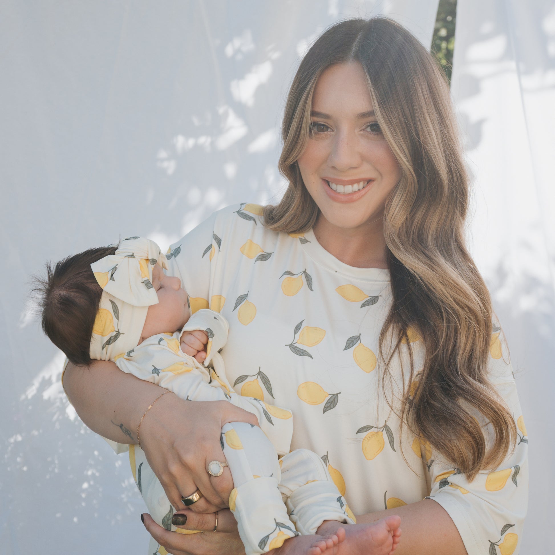 A happy woman with long, wavy hair holds her baby. Both wear forever french baby Bamboo Zip Pajamas in lemon print, paired with a matching headband, set against a softly lit white fabric and greenery backdrop.