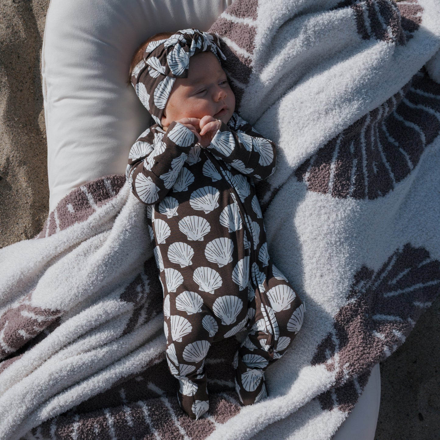 A baby wears forever french baby's Bamboo Zip Pajamas | Seashells—breathable sleepwear with a matching headband—while sleeping on a cushion, wrapped in a cozy blanket, all featuring the white seashell design.