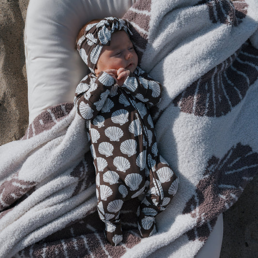 A baby wears forever french baby's Bamboo Zip Pajamas | Seashells—breathable sleepwear with a matching headband—while sleeping on a cushion, wrapped in a cozy blanket, all featuring the white seashell design.