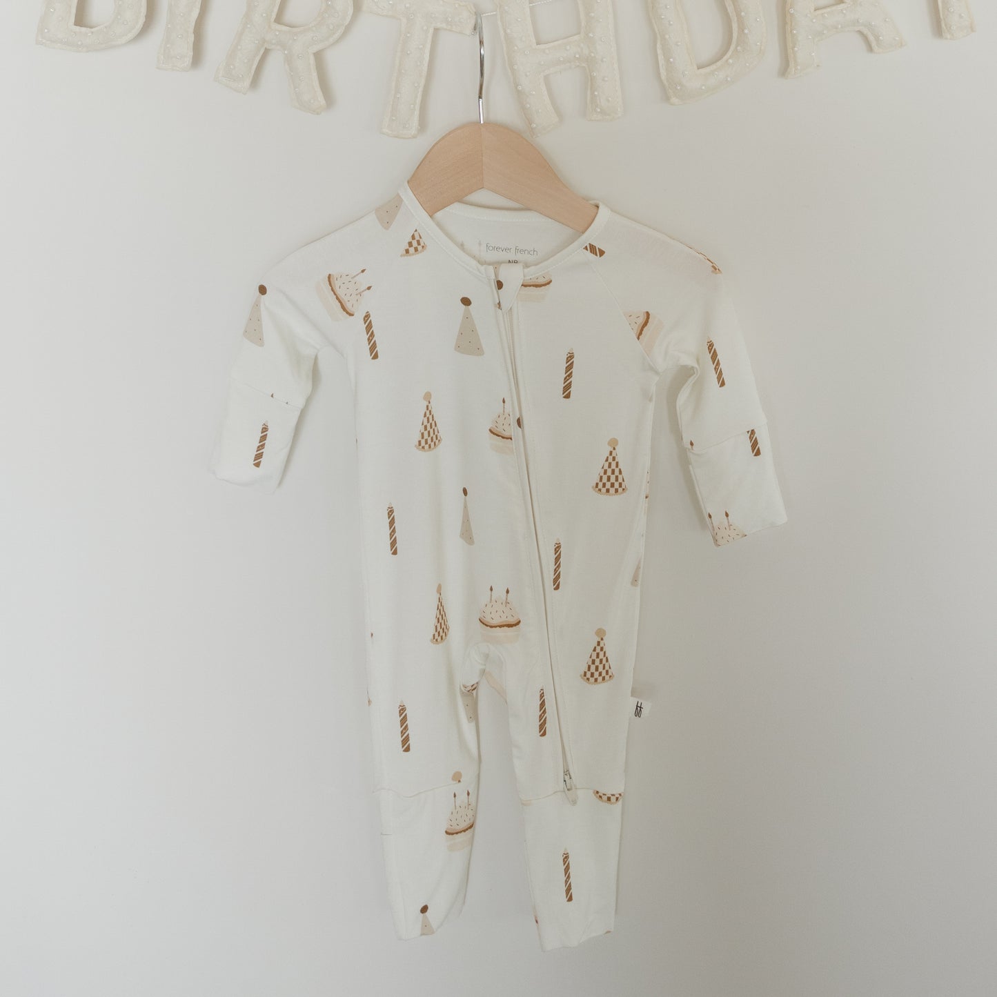 A pair of white "Bamboo Zip Pajamas | Birthday Boy" by forever french baby, made from hypoallergenic bamboo fabric and featuring birthday-themed prints like cake and candles, hangs on a wall beneath a cream-colored "BIRTHDAY" banner.